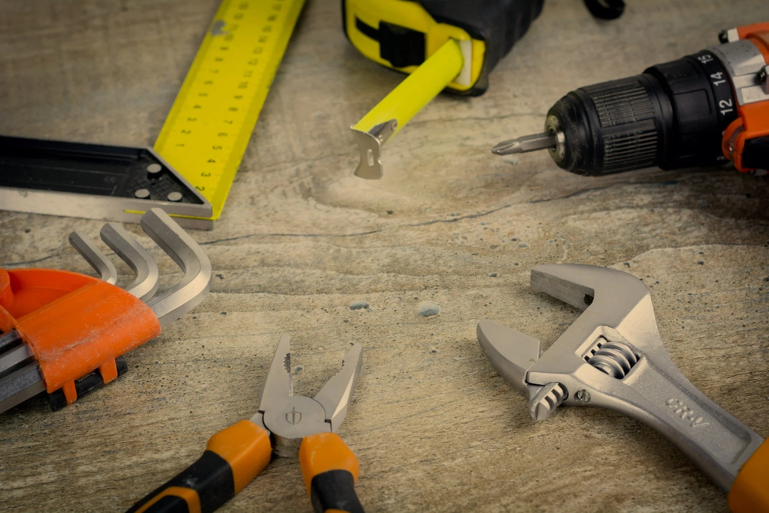 Various tools on a workbench, including a cordless drill, adjustable wrenches, needle nose pliers, a measuring tape, and a utility knife.