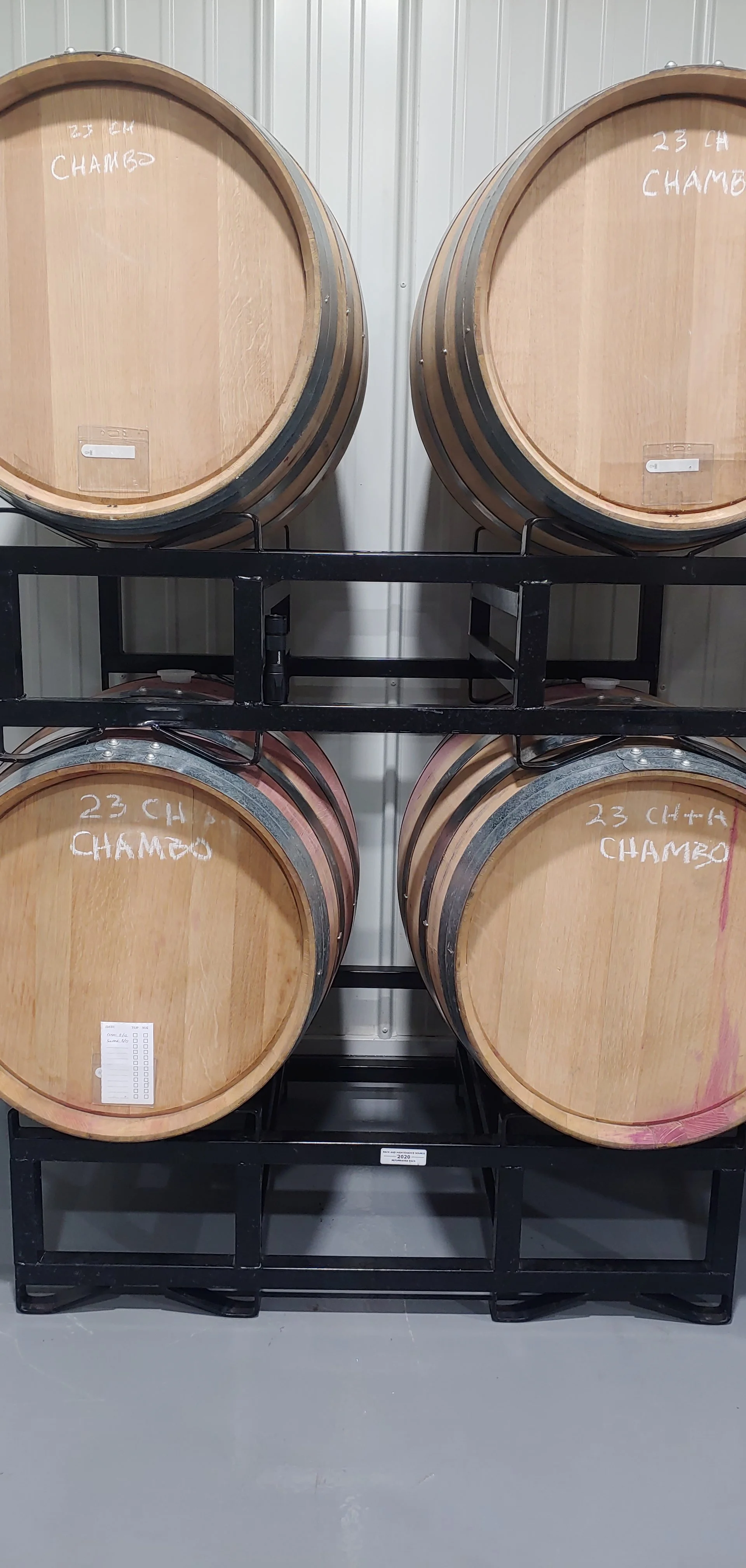 Four wooden barrels labeled '23 CH CHAMBO' stacked on a black metal rack in a storage room.