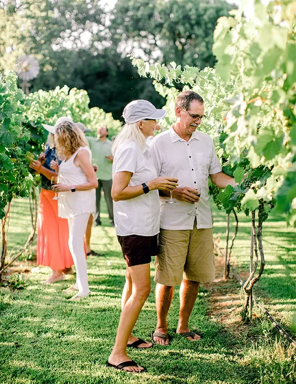 Group of people enjoying wine tasting in an Oklahoma vineyard on a sunny day, with green grapevines and trees in the background.