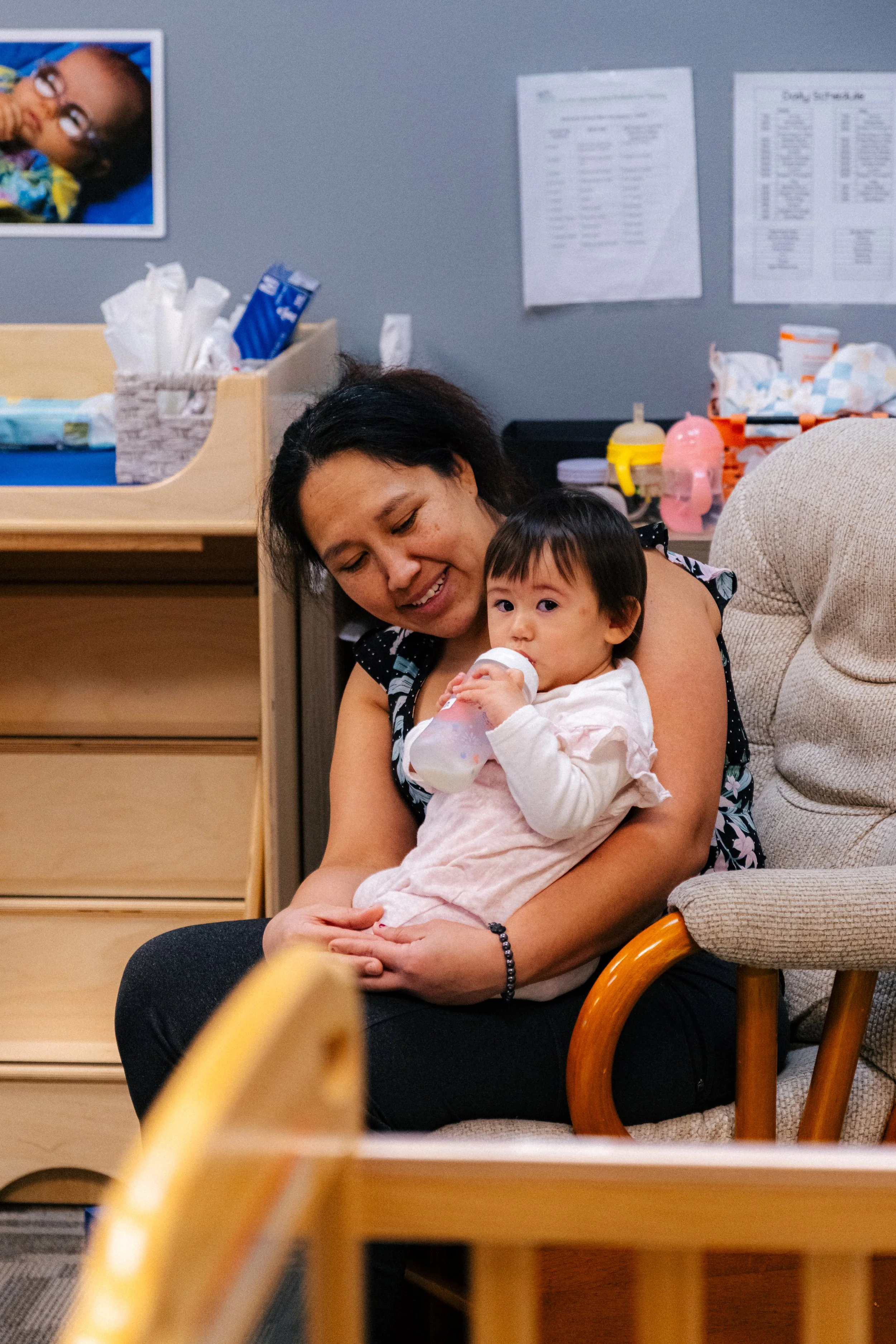 A woman holding a young child drinking from a bottle in a waiting room or medical office, with various items and posters on the wall behind them.