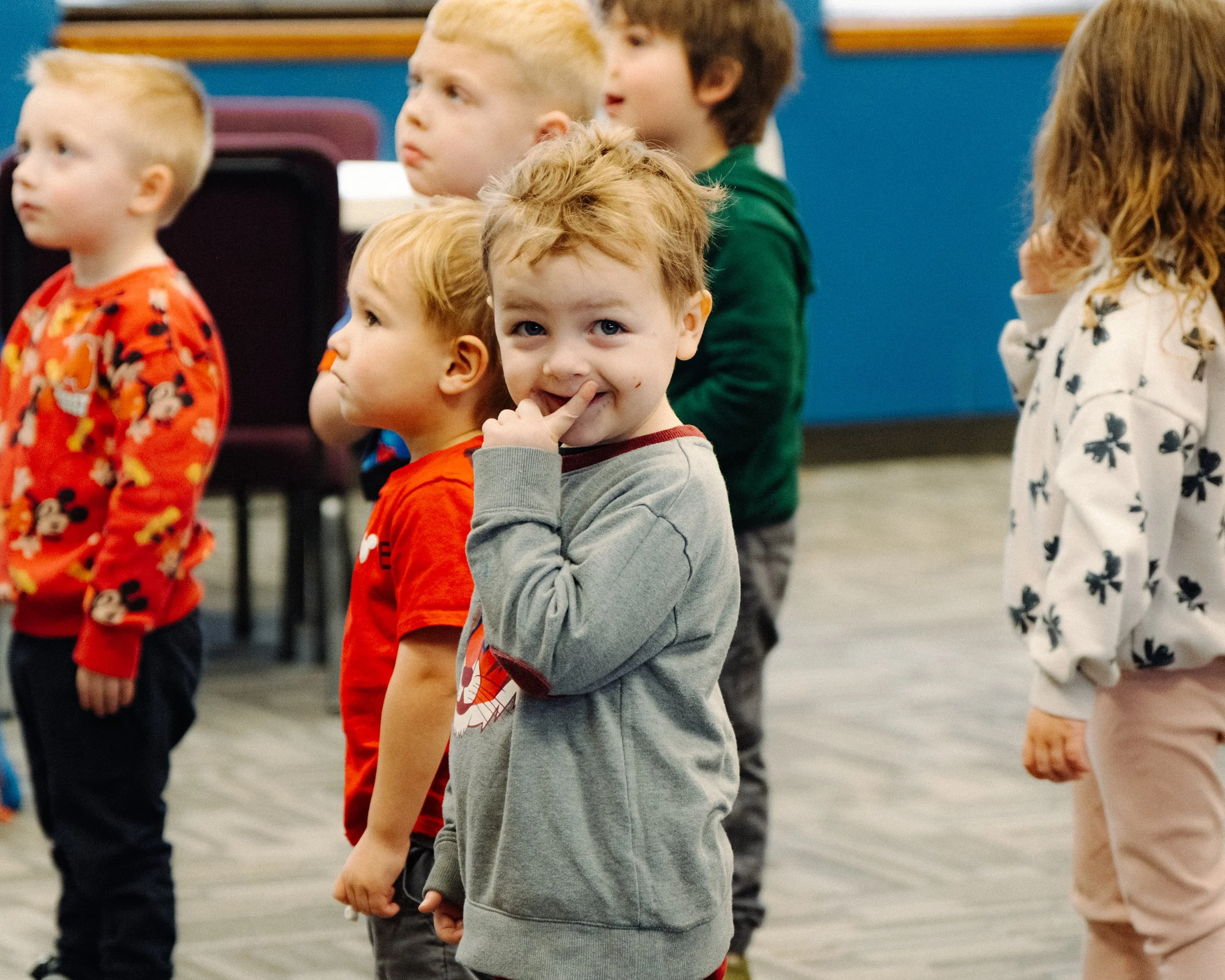 A group of young children standing in a line indoors, some looking forward, one child in the foreground smiling and finger near mouth.