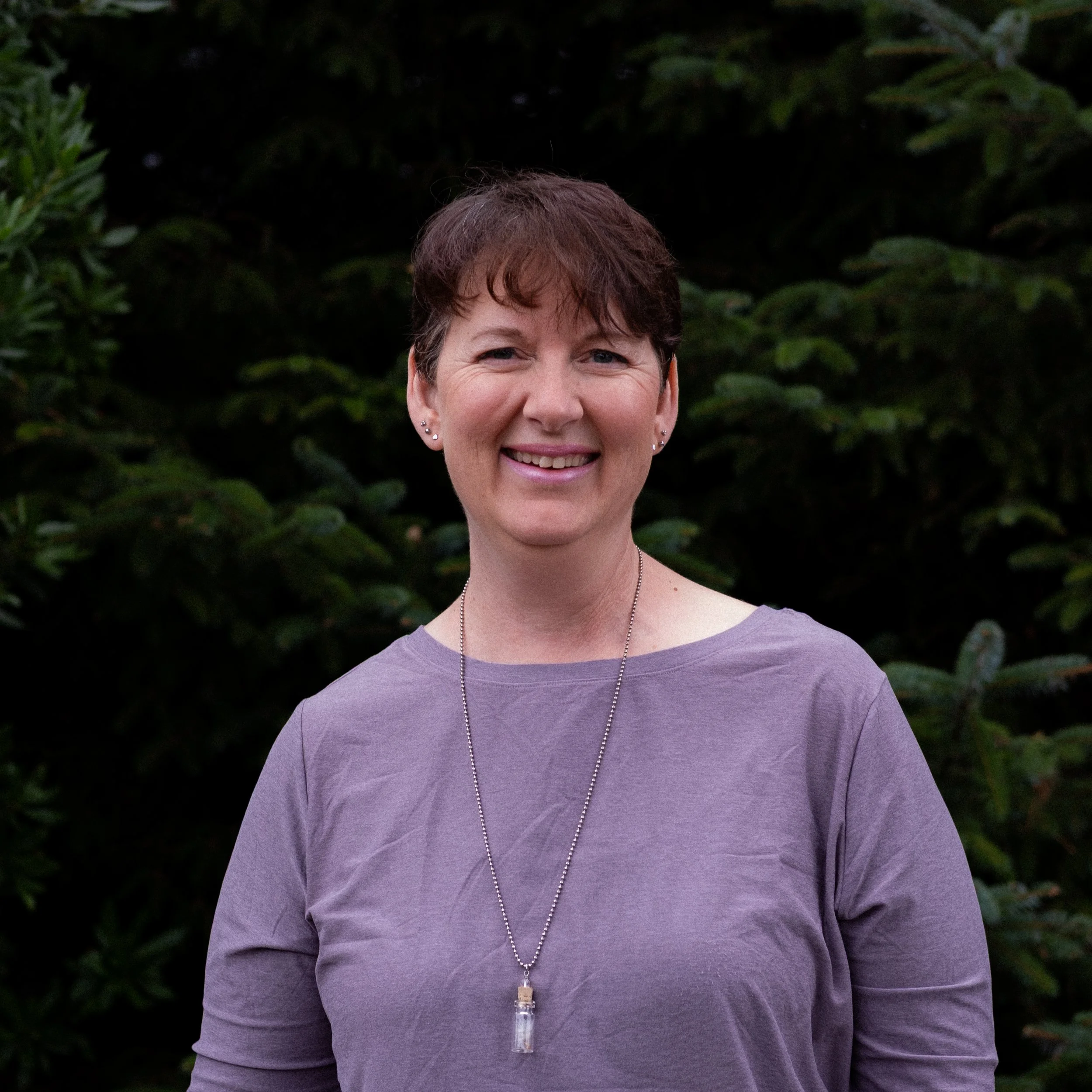 A woman with short brown hair, wearing a purple shirt and a necklace, smiling outdoors in front of greenery.