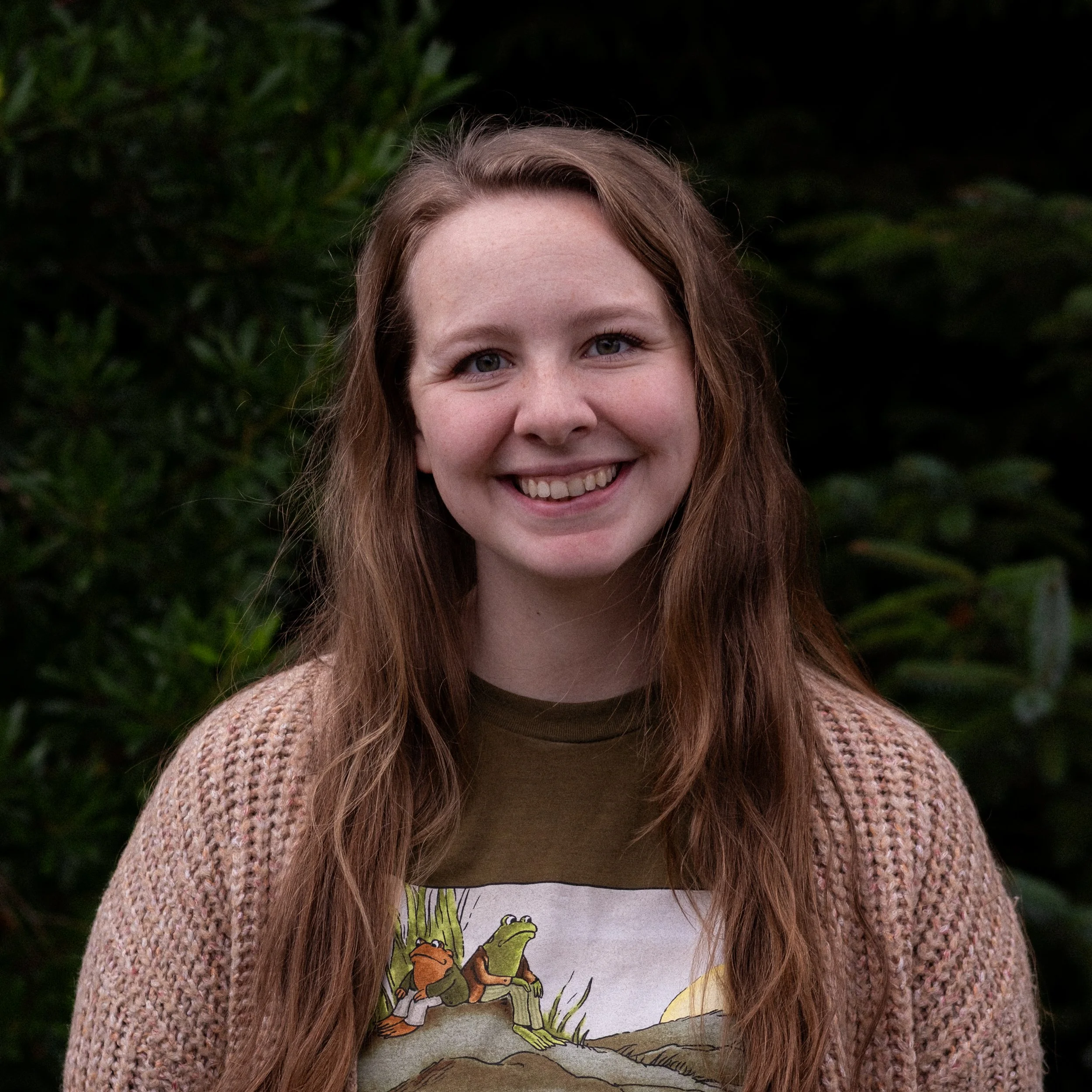 A young woman with long, wavy red hair smiling outdoors with green foliage in the background, wearing a beige knitted cardigan over a graphic T-shirt.