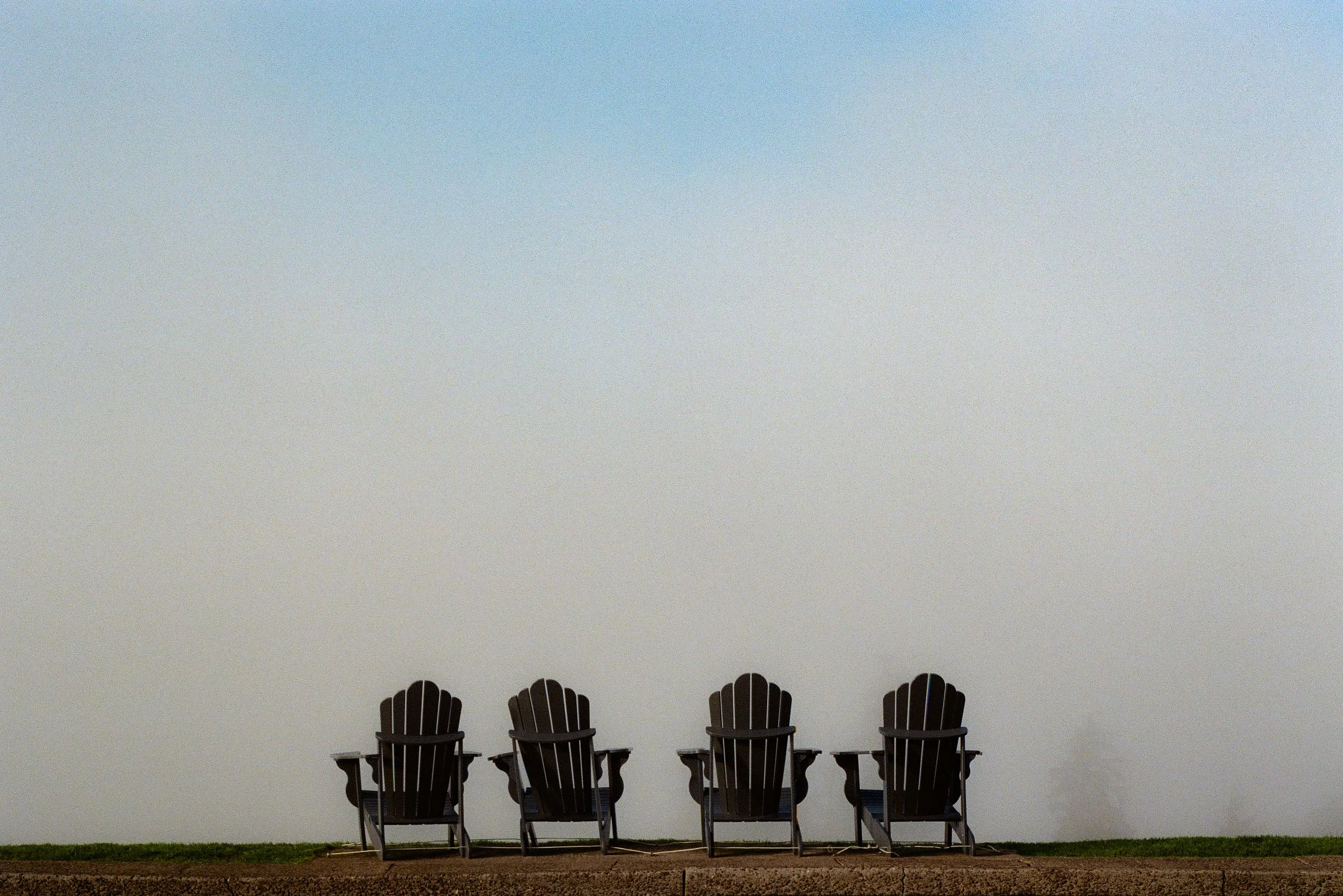 Four empty Adirondack chairs facing a clear sky, with a grassy and dirt ground in the foreground.