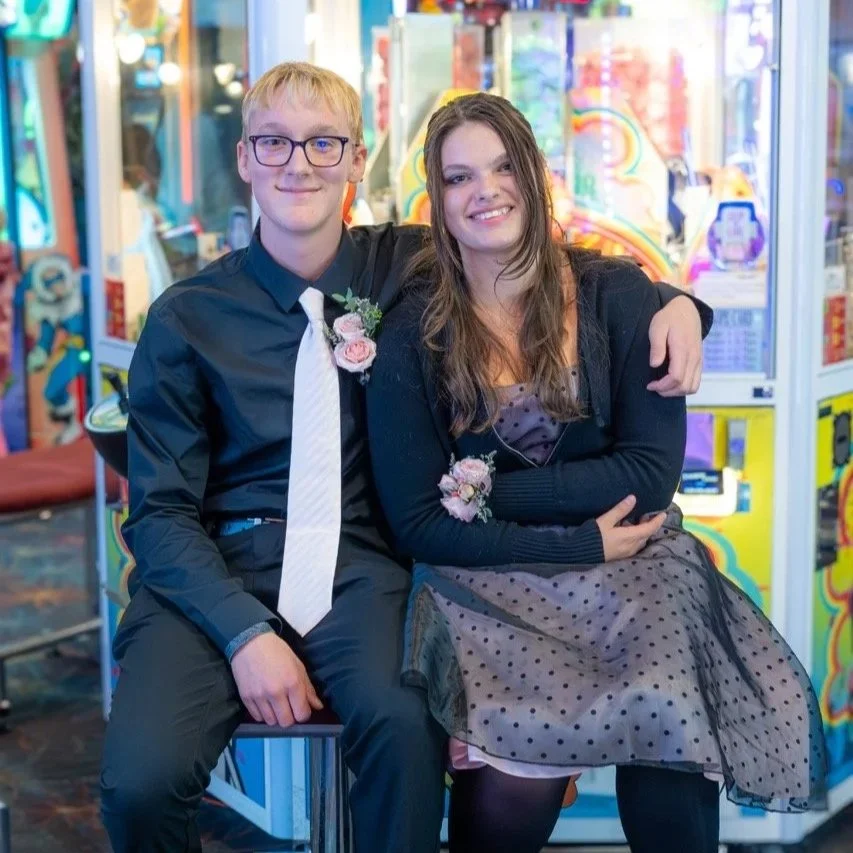 A young man and woman dressed formally, sitting together in an arcade, smiling. The young man has blond hair, glasses, and is wearing a black shirt with a white tie and corsage. The young woman has long brown hair, is wearing a black dress with a she