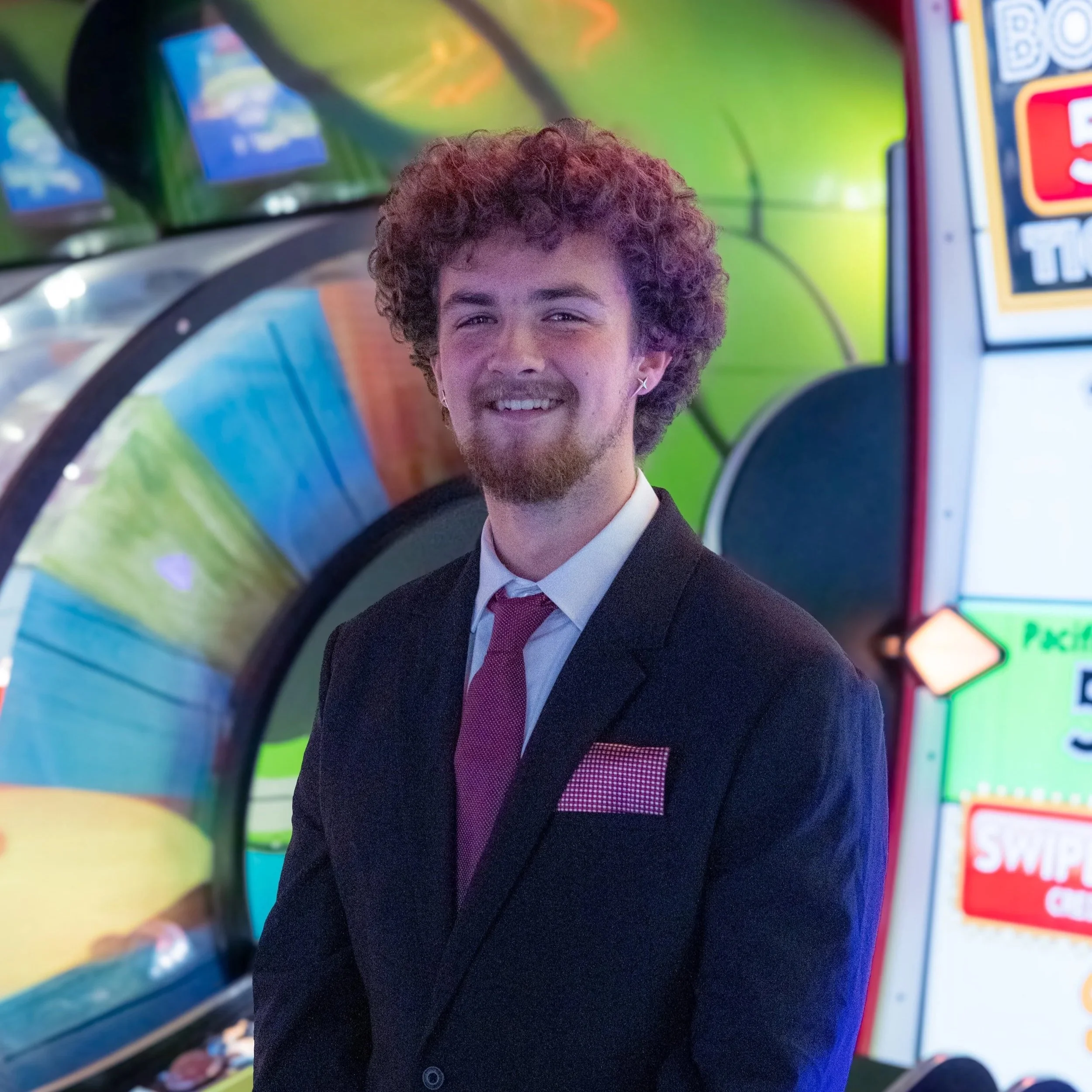 Young man with curly hair, beard, and earrings smiling in front of colorful arcade game machines.