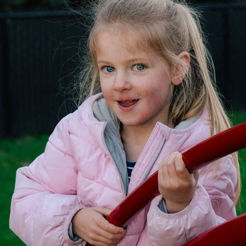 A young girl with blonde hair and blue eyes, wearing a pink jacket, holding a red slide on a playground, outdoors with grass and a black fence in the background.
