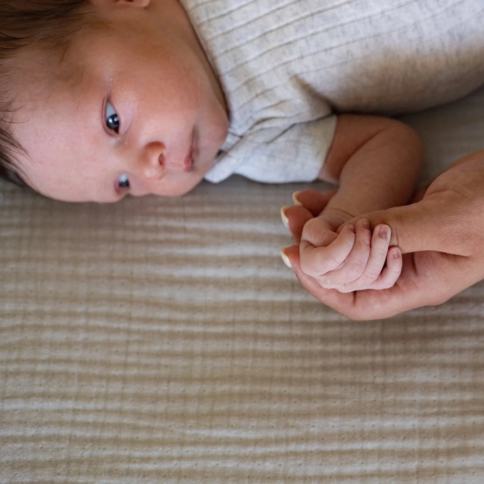 A baby lying on a beige striped surface holding an adult's finger.