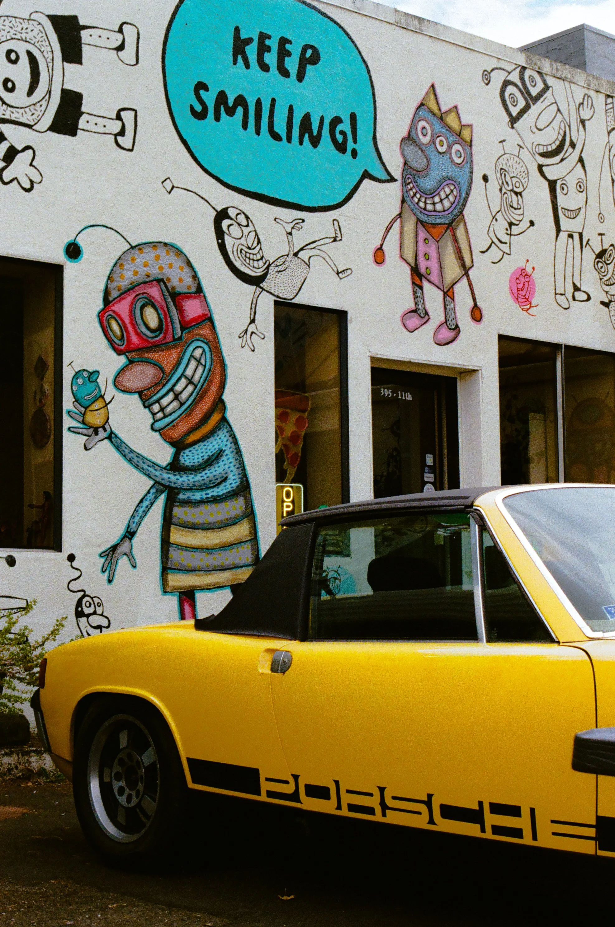 A vibrant mural featuring cartoon characters and a speech bubble that says 'Keep Smiling!' next to a vintage yellow Porsche convertible parked in front of the wall.