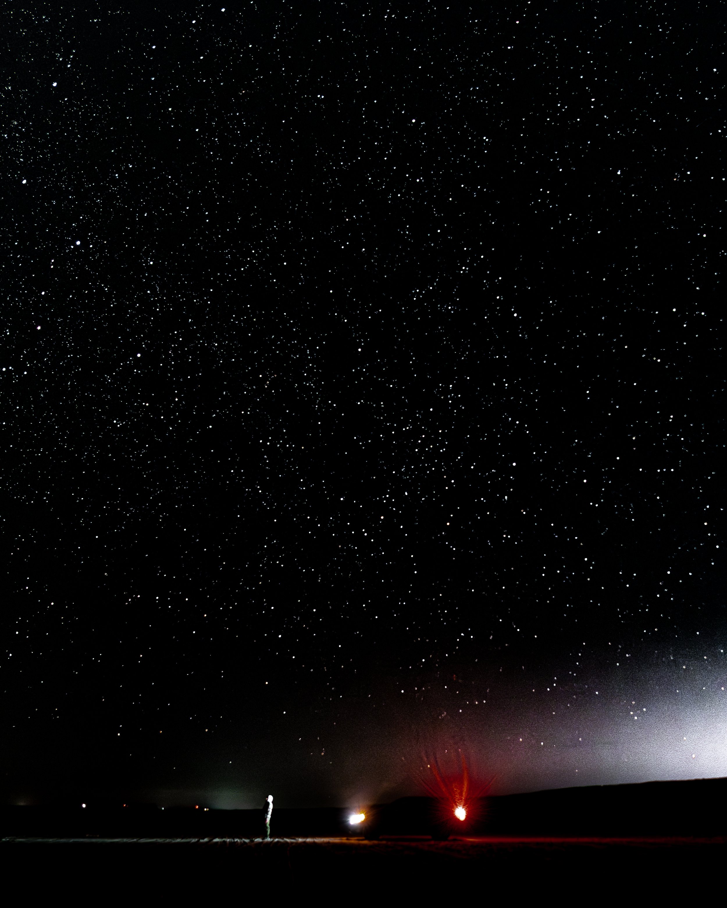 Night sky filled with stars, with a person standing on a flat surface, and some lights or small structures on the horizon.
