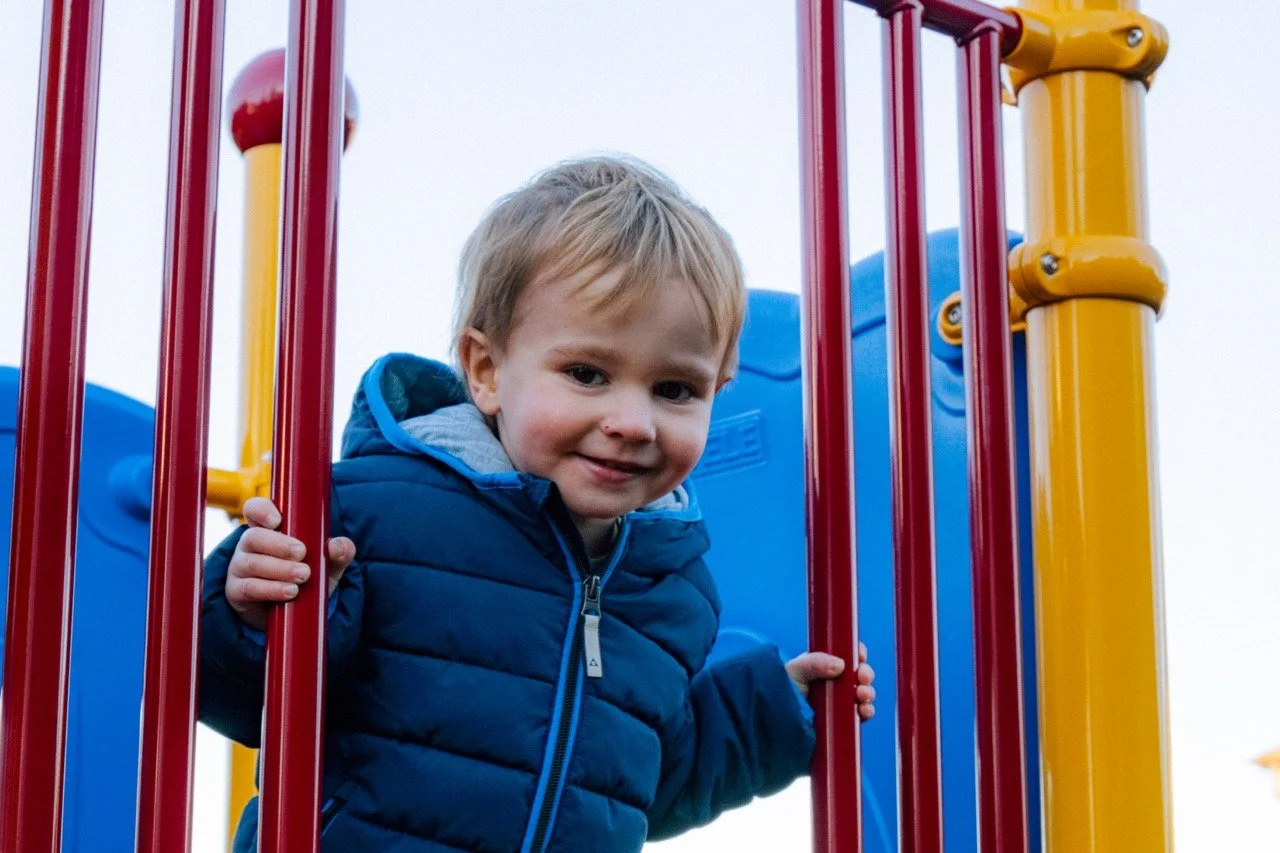 A young boy with blonde hair and a blue jacket smiling while playing on a colorful playground.