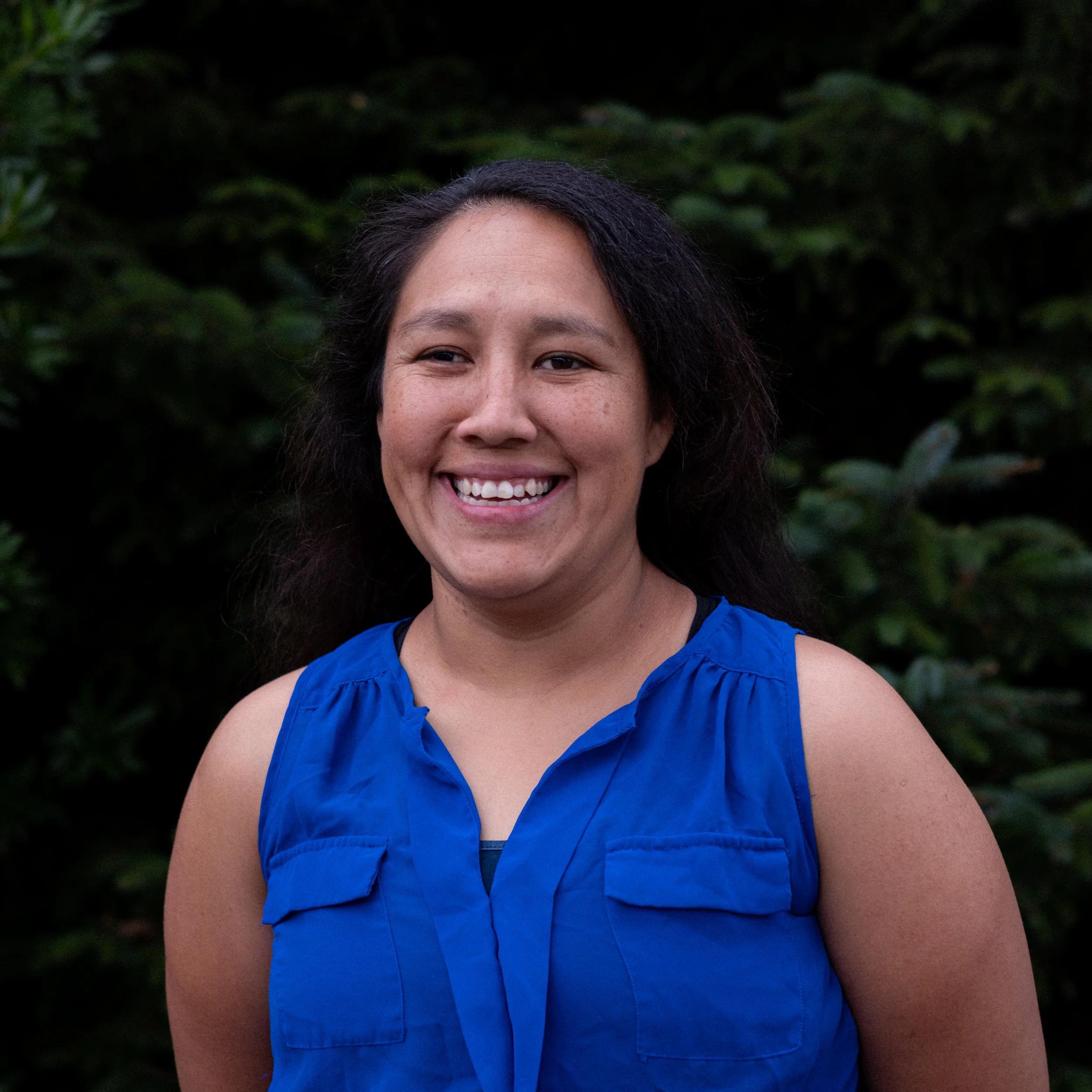 A woman with long dark hair smiling outdoors, wearing a blue sleeveless top, with green foliage in the background.