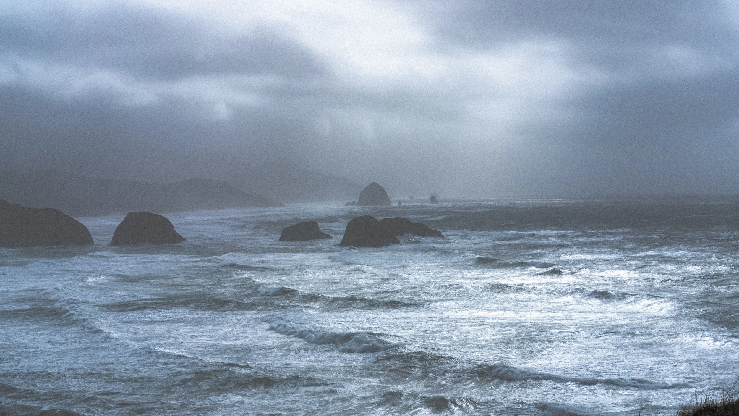 Stormy ocean scene with large rocks in the water, overcast sky, and distant landforms in the background.