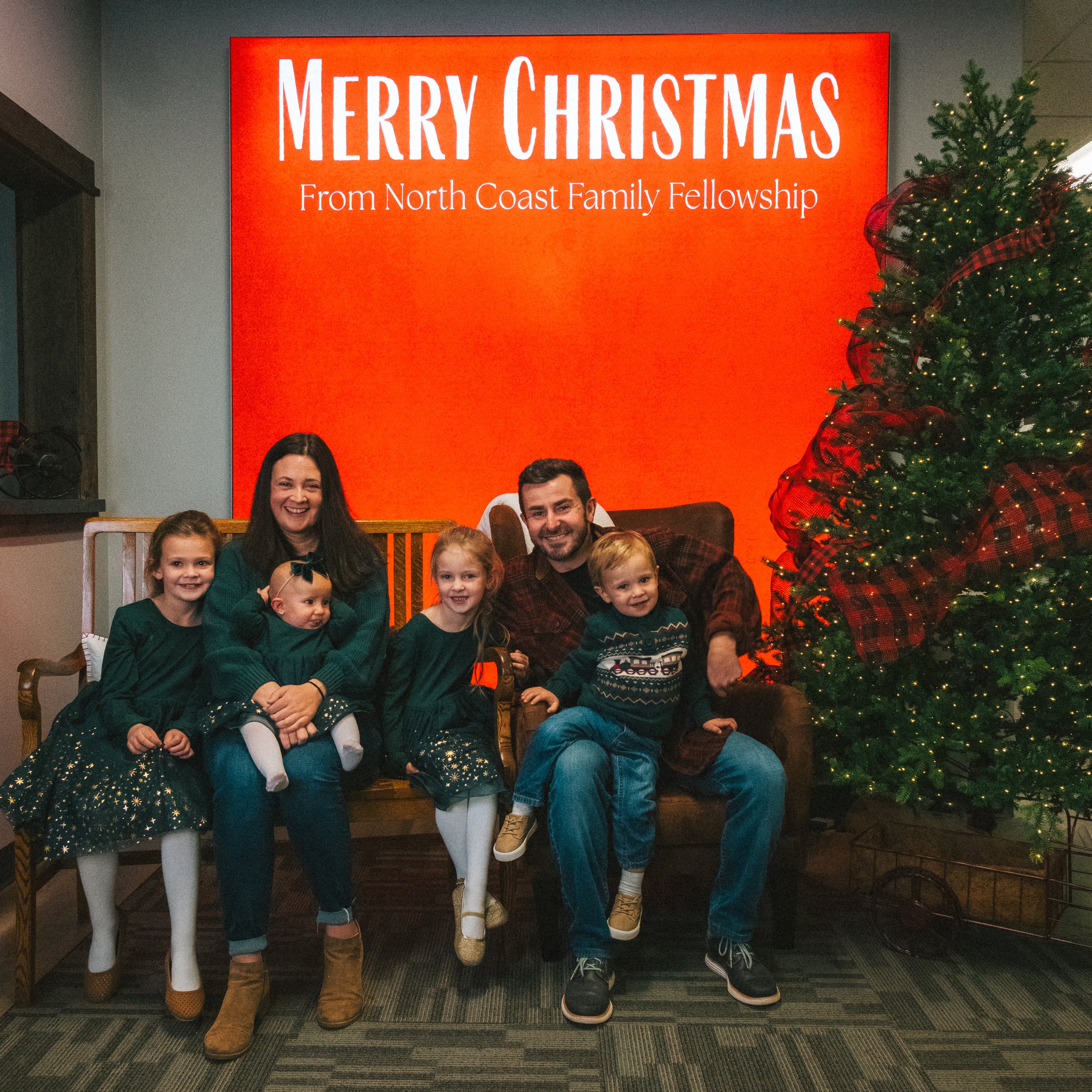 Family of five sitting on a bench in front of a large red sign that says 'Merry Christmas from North Coast Family Fellowship', next to a decorated Christmas tree.