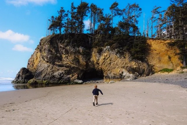 A person walking alone on a sandy beach with a large rocky cliff covered in trees in the background under a blue sky.
