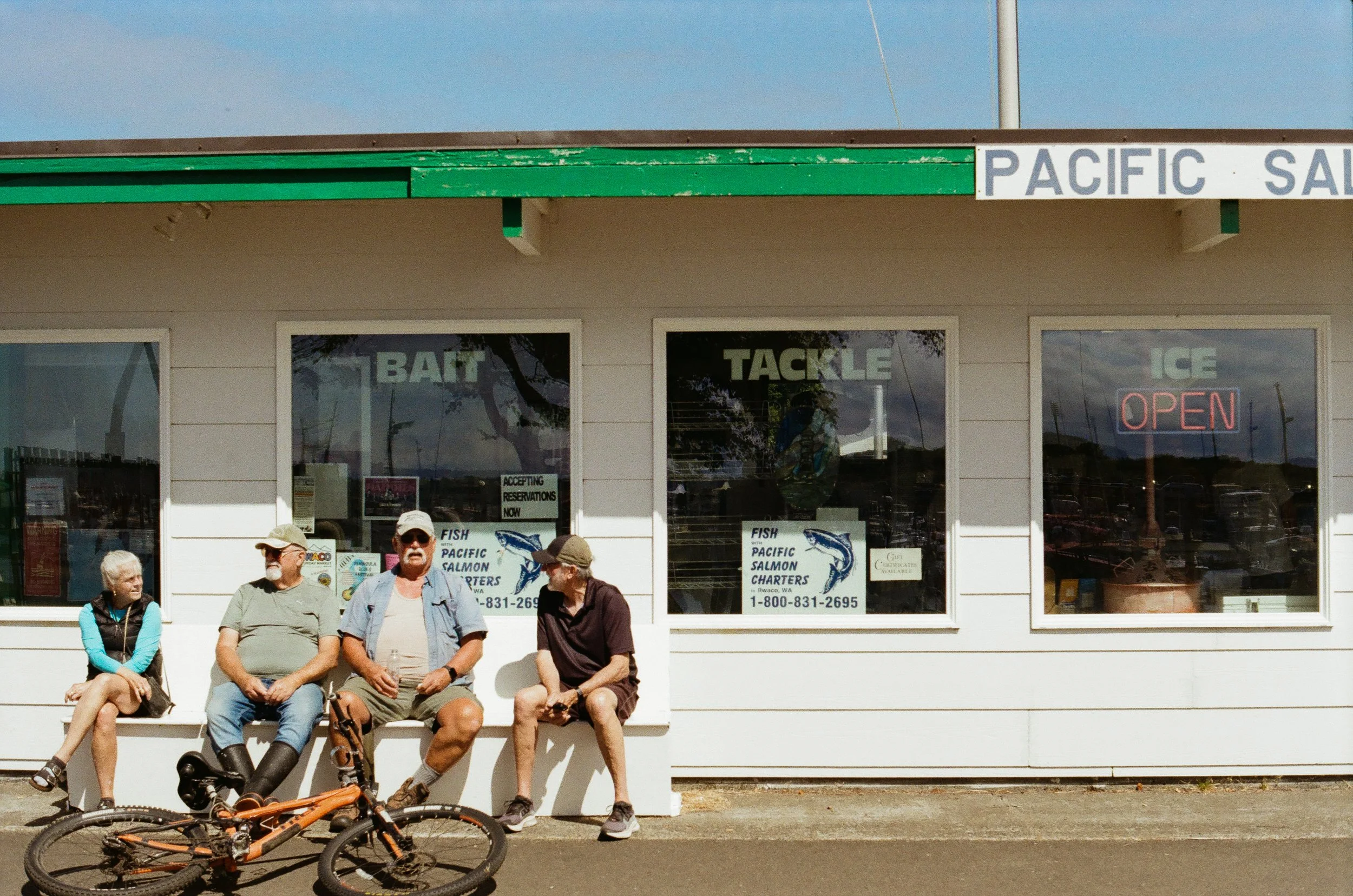 Four people sitting on a white bench outside a bait shop, with a bicycle in front. The shop has large windows with signs for fish, Pacific salmon, and charters, and an illuminated 'Open' sign.