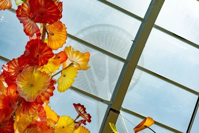 Colorful glass flower installation viewed from below, with the Seattle Space Needle visible through glass ceiling.