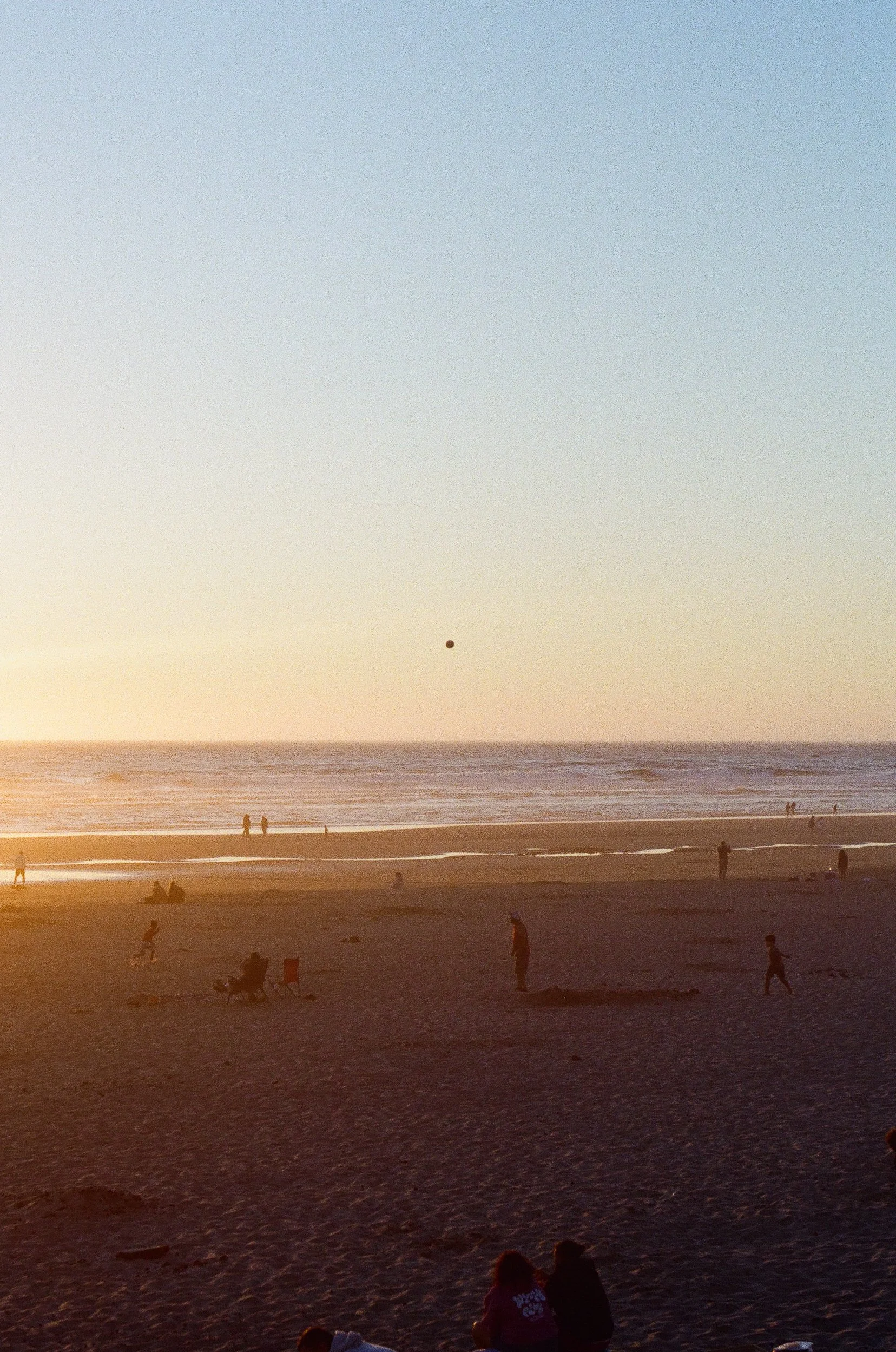People enjoying the beach at sunset, some sitting, some walking, with the ocean waves and a clear sky in the background.