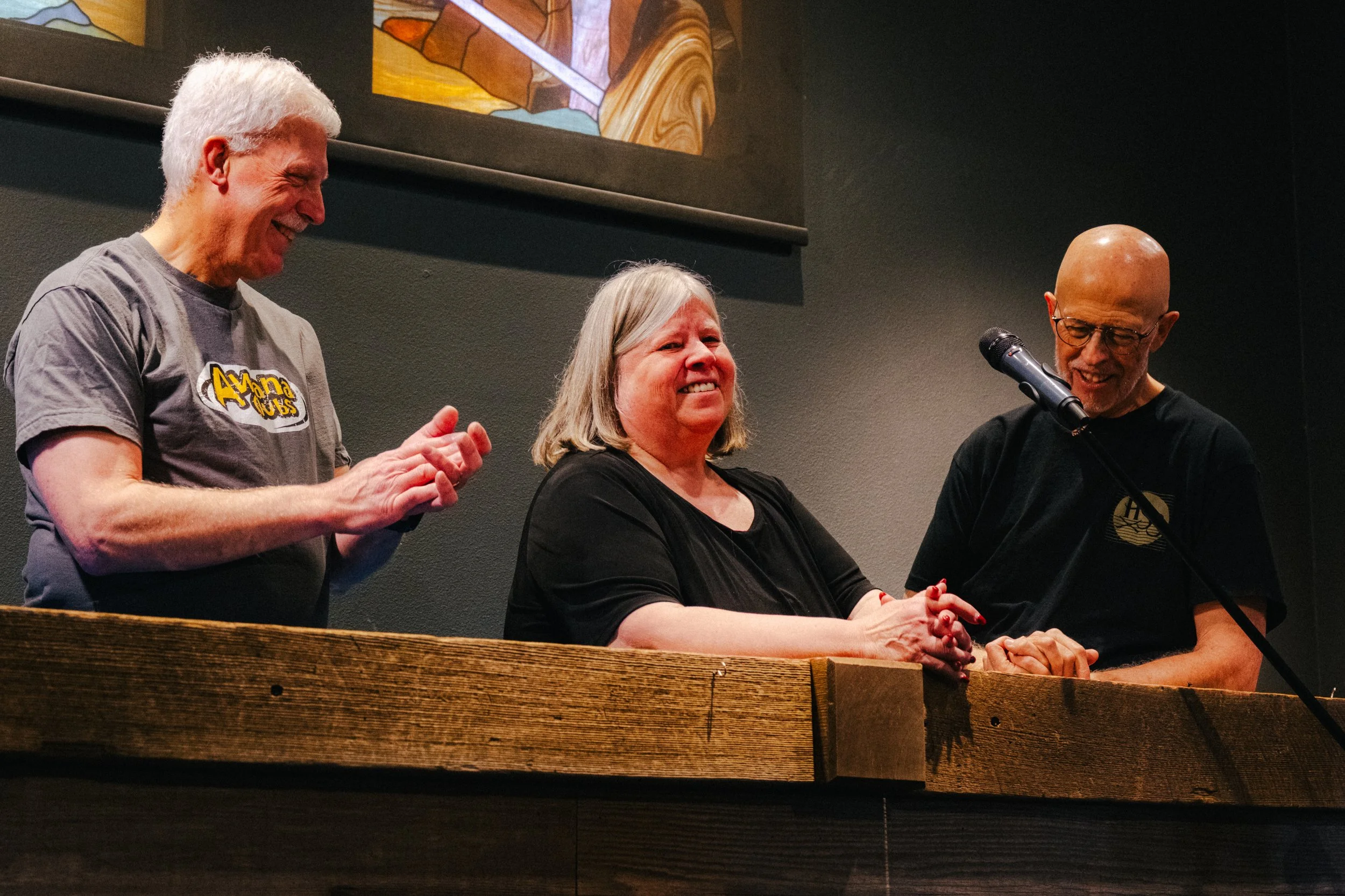 Three people standing behind a wooden podium, with one woman in the middle appearing emotional and tearing up, while the two men on either side are smiling and looking at her. The man on the left has gray hair and is wearing a gray T-shirt with a log
