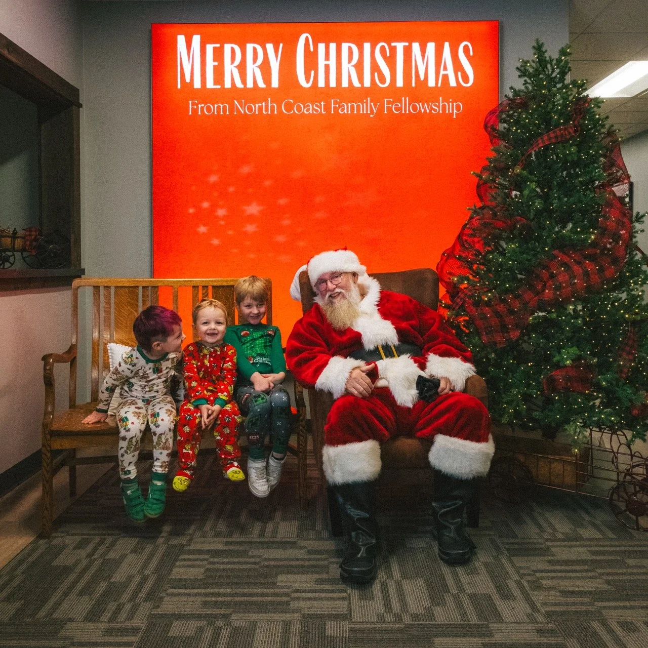 Santa Claus sitting with three children on a bench, next to a decorated Christmas tree, with a large red sign behind them that reads 'Merry Christmas from North Coast Family Fellowship'.