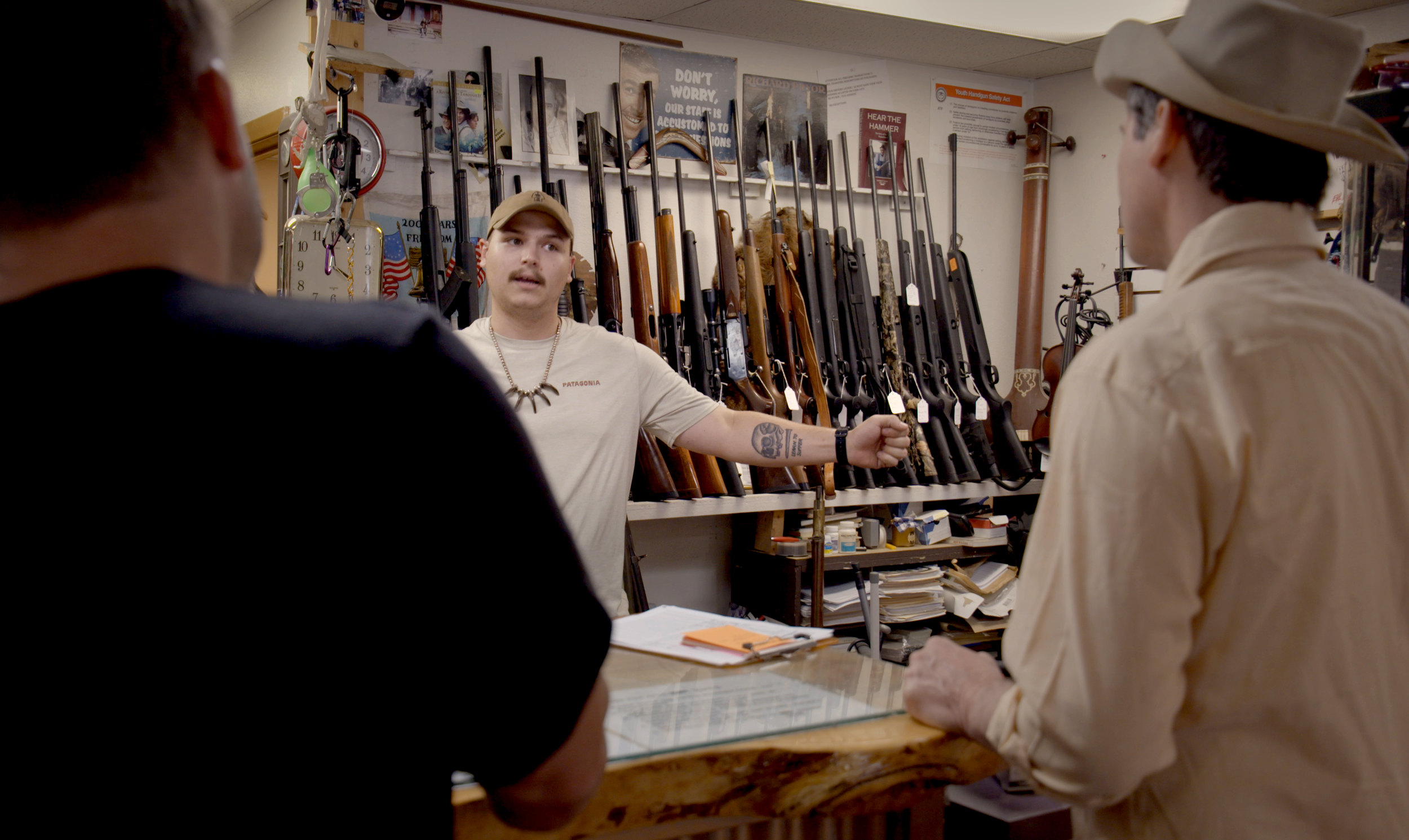 Three men stand at a gun store counter with a wall of rifles behind them. The man in the middle is gesturing with his hand and speaking, while the other two listen.