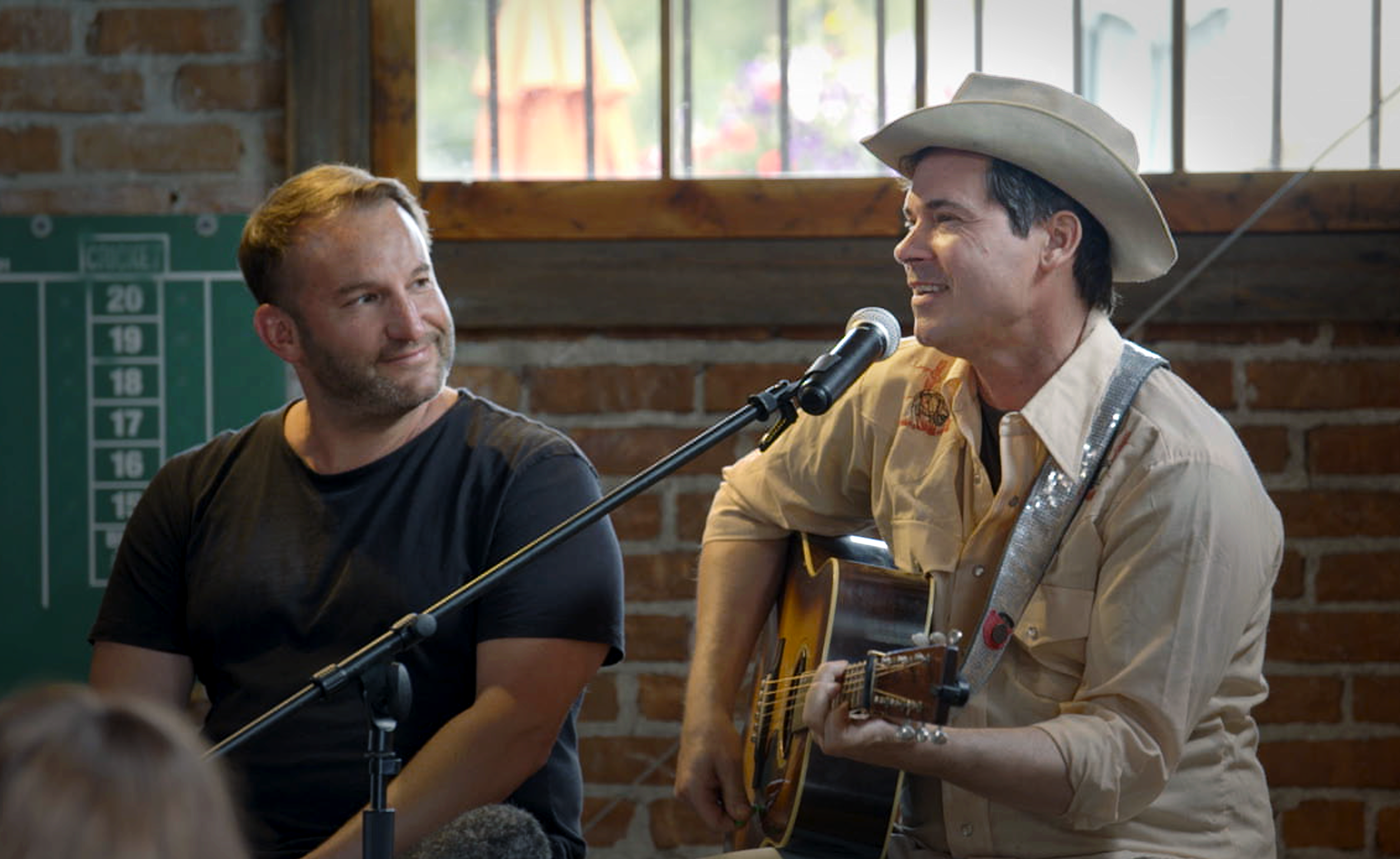 Two men in a rustic brick-walled room, one playing an acoustic guitar and singing into a microphone, the other listening and smiling, with a window in the background.