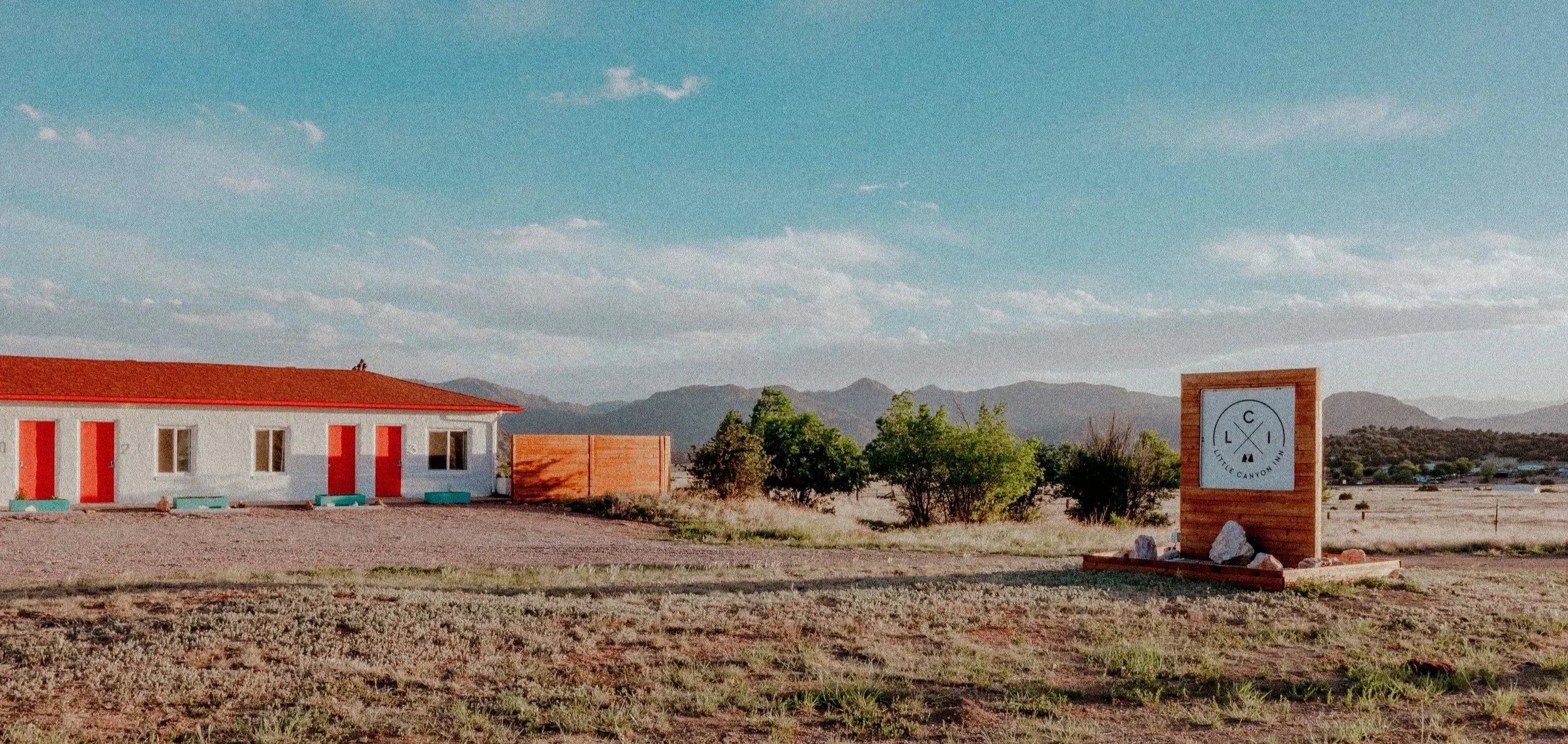 A white building with a red roof and red doors, yellowish gravel ground, trees, a wooden sign that reads 'Little Canyon Inn' with mountains in the background under a partly cloudy sky.