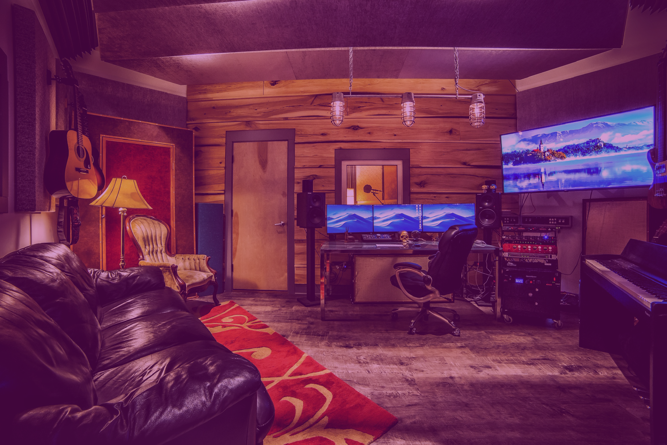 A music and recording studio with a leather sofa, vintage armchair, three computer monitors, large wall-mounted TV, keyboard, and various music equipment, all set in a cozy room with wood-paneled walls and ceiling.