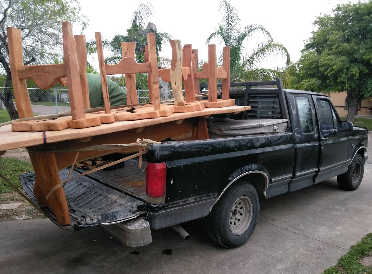 A black pickup truck parked outdoors with a wooden table and chairs loaded in the bed of the truck. The chairs are stacked upside down on top of the table.