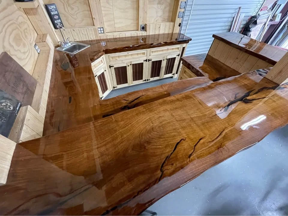 A wooden bar with a glossy finish in a woodworking shop, showing detailed wood grain and natural cracks, with a small sink on the left and cabinets underneath.