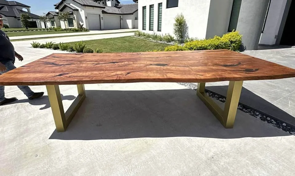 A wooden outdoor table with a polished surface and gold-colored metal legs, situated on a concrete patio in front of a modern house with landscaping.