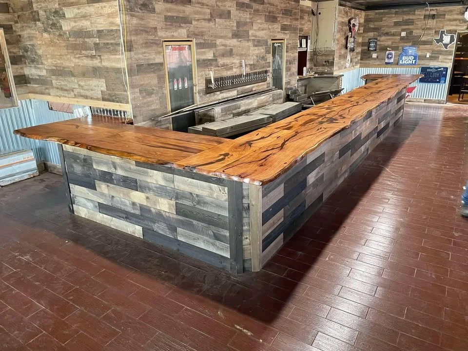 Long wooden bar with a polished natural wood top and a rustic wood and metal panel base inside a restaurant or pub.
