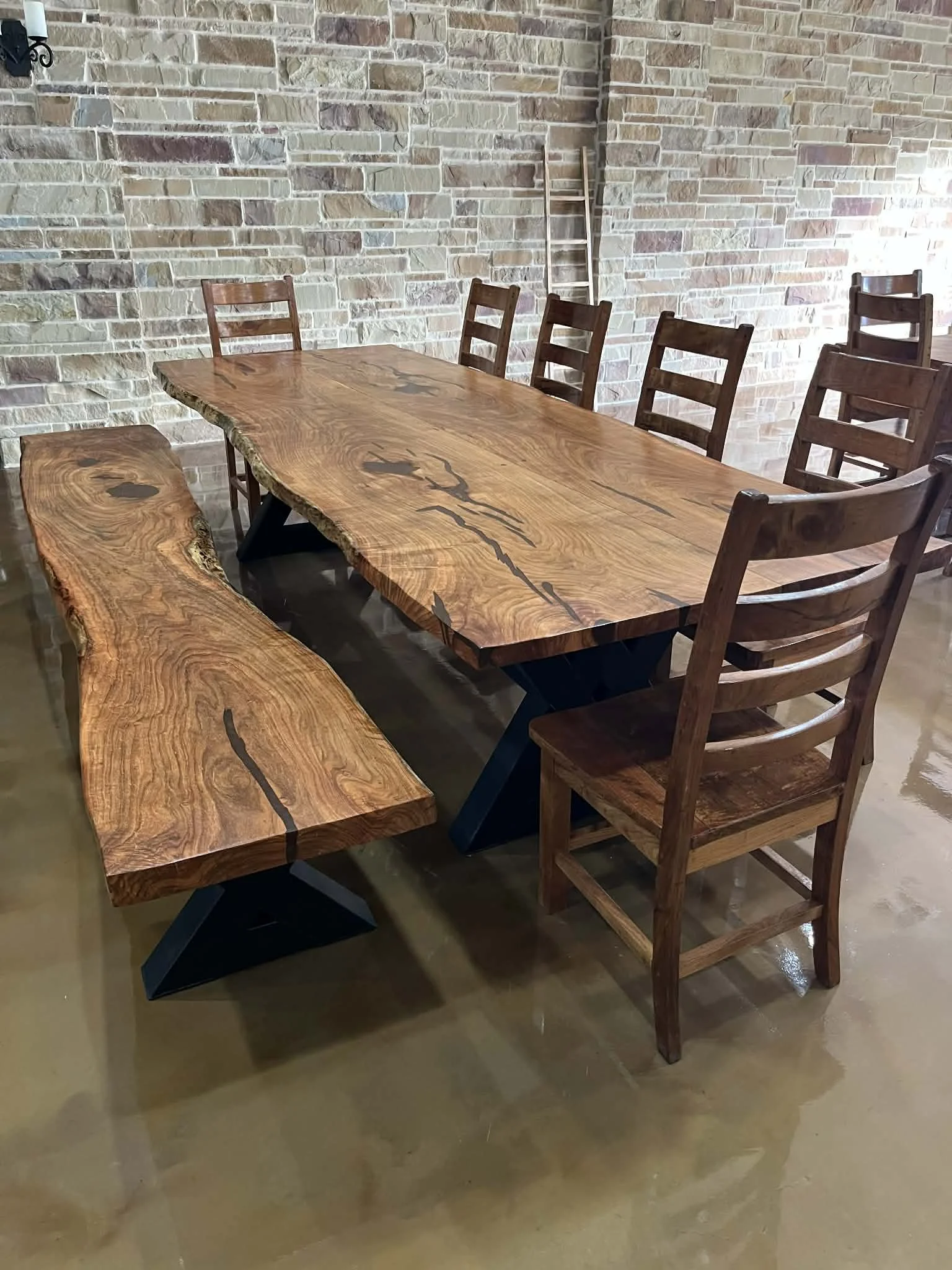 A rustic dining table made of thick, natural wood with a live edge, accompanied by matching wooden chairs and a bench, set against a brick wall.