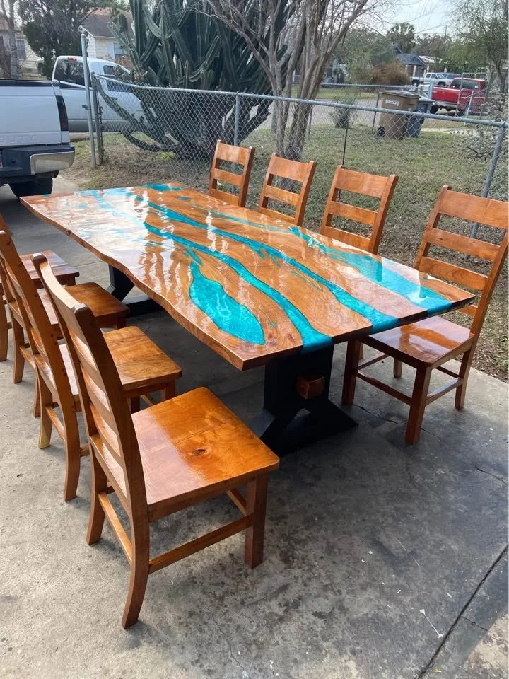 A wooden dining table with a unique epoxy design in blue and natural wood finish, surrounded by six matching wooden chairs, outdoors on a concrete patio.