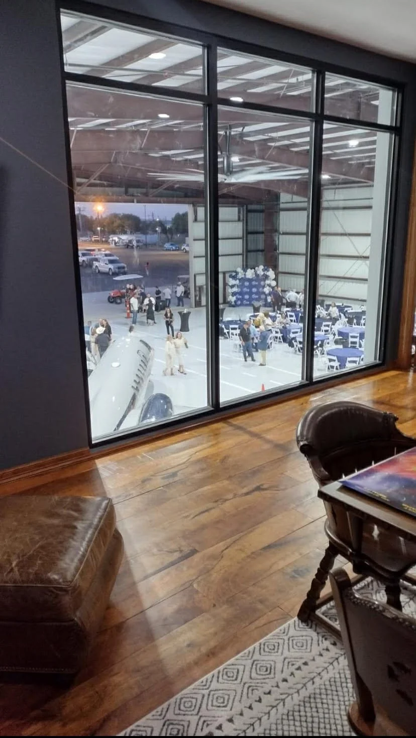 View of a social event or gathering inside a large shed or open building seen through a large window. There are tables with blue tablecloths, people mingling outside, and balloons in the background.