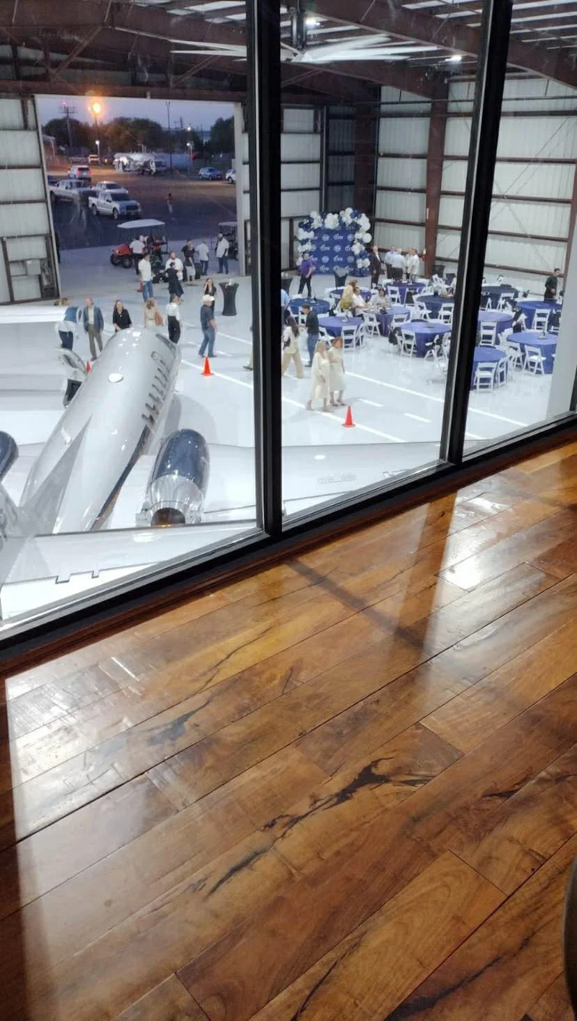 View from inside a hangar looking out onto an event space with a vintage airplane parked outside, decorated with balloons and tables set up for a celebration, with people mingling and a person on a golf cart nearby.