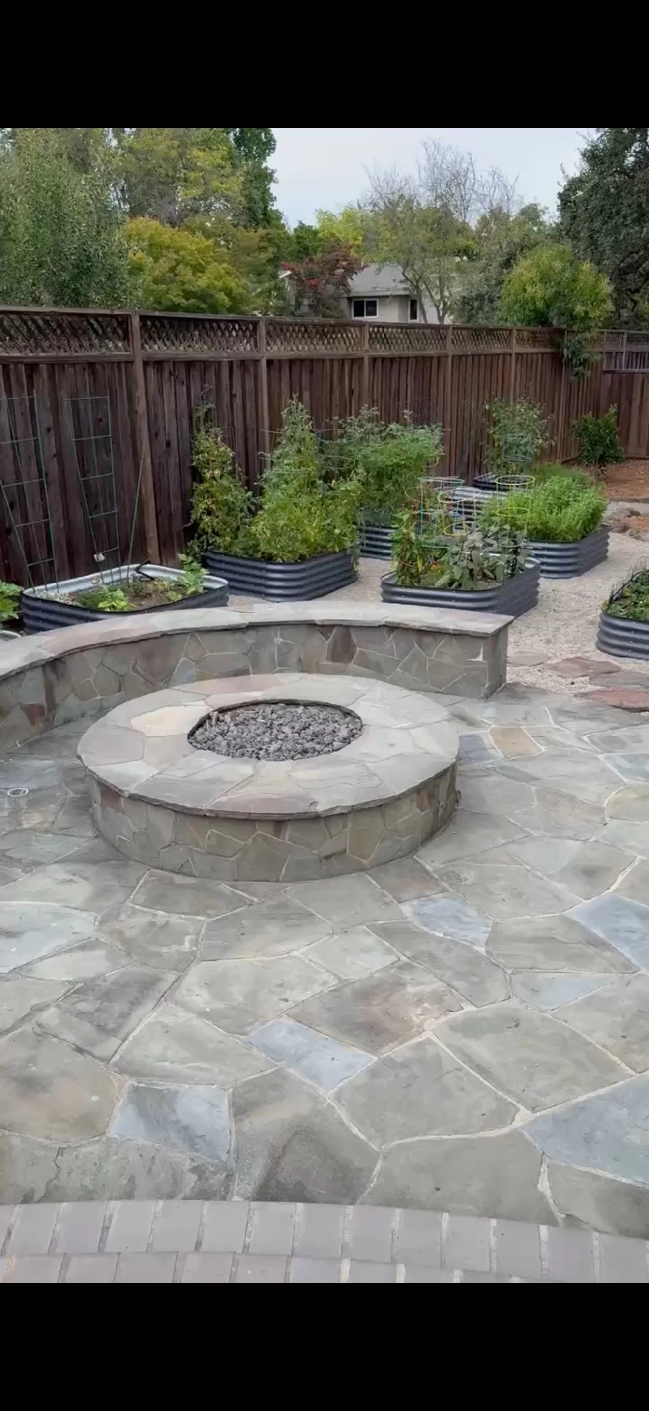 Backyard with stone patio, fire pit, and raised garden beds with plants, enclosed by wooden fence.