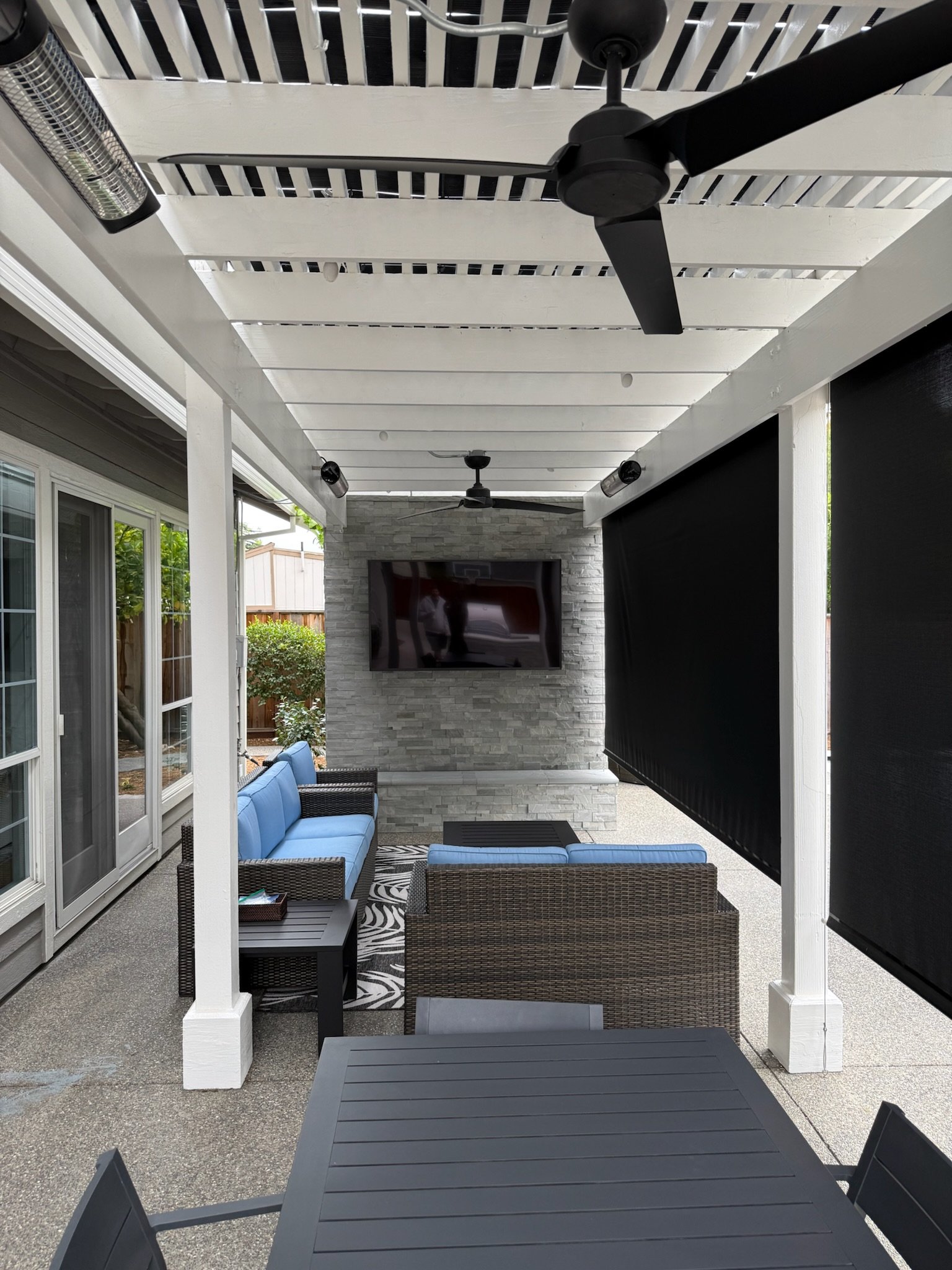 A covered outdoor patio area with a black dining table, outdoor rattan furniture with blue cushions, a flat-screen TV mounted on a stone wall, ceiling fans, and black retractable blinds.