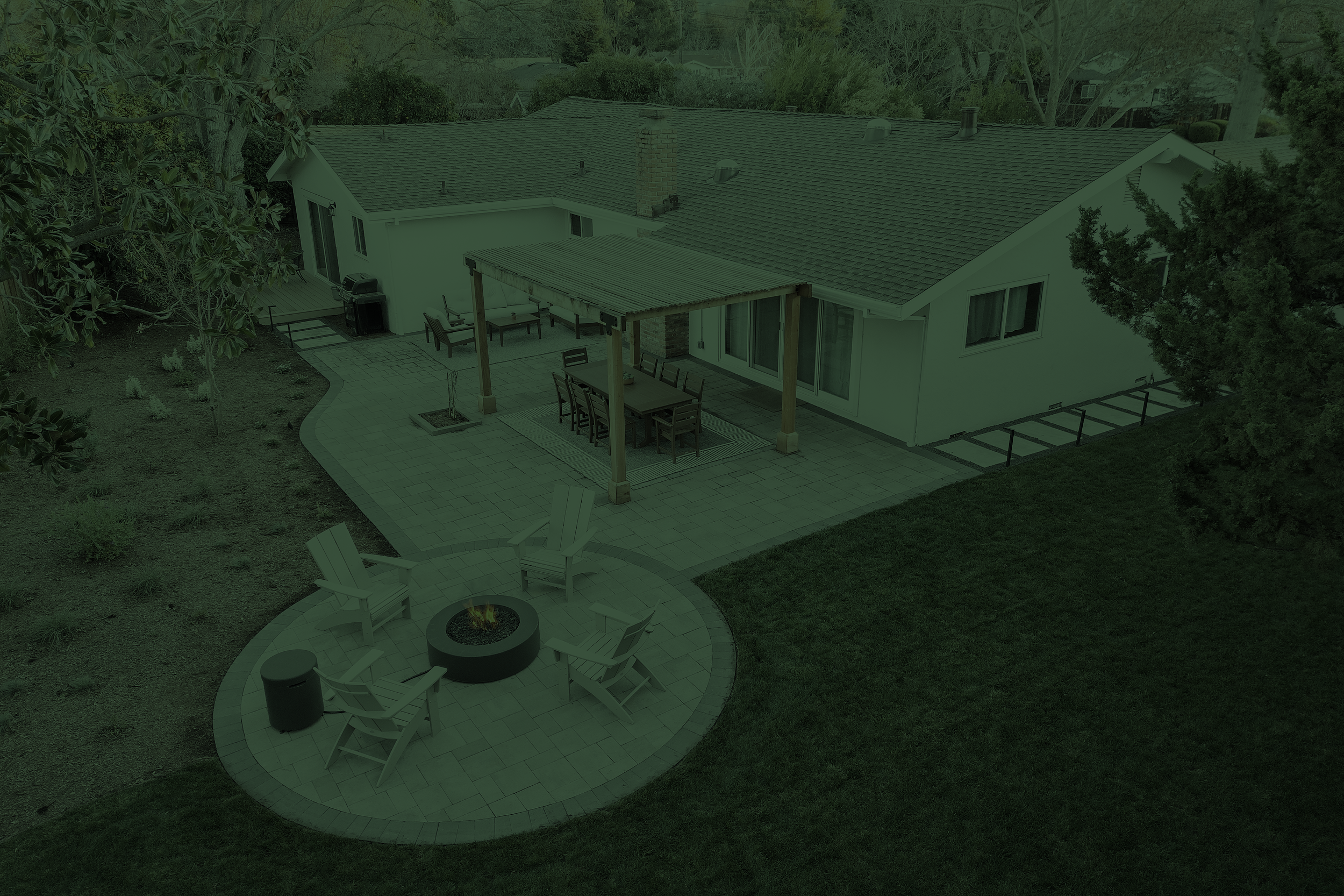 Aerial view of a backyard patio with outdoor furniture, a fire pit, and a house with a tiled roof surrounded by trees and greenery.