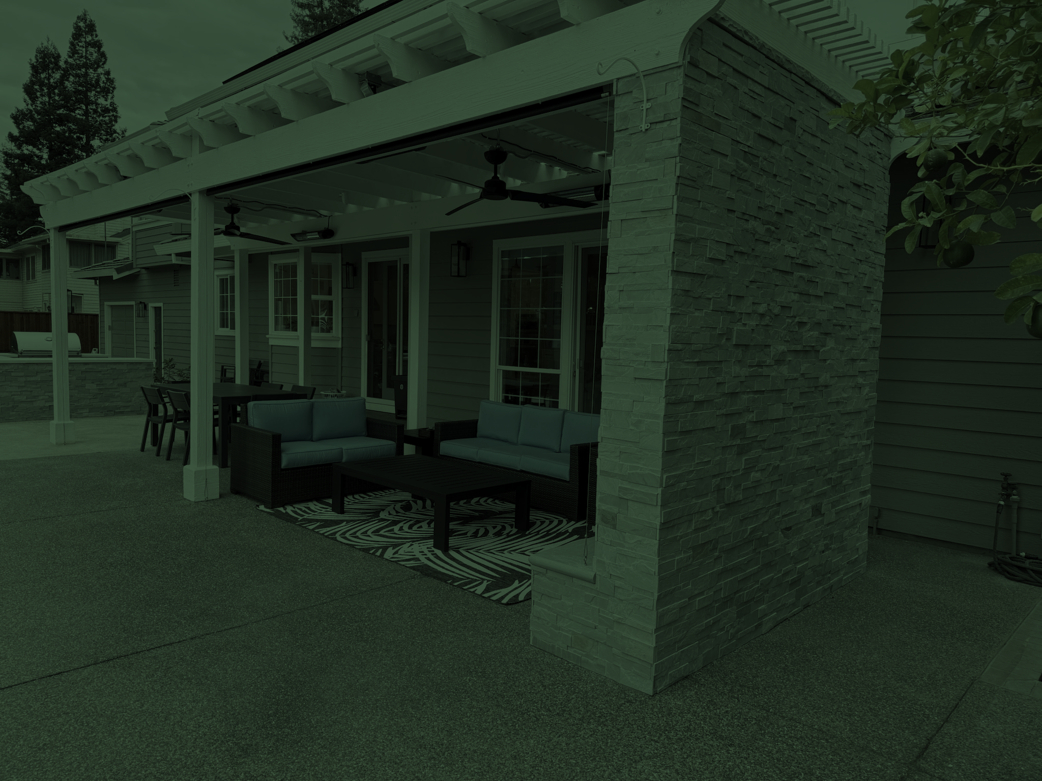 Backyard patio area with outdoor furniture, a grill, and a covered seating space with ceiling fans. There is a brick pillar and a pergola structure, with trees visible in the background.