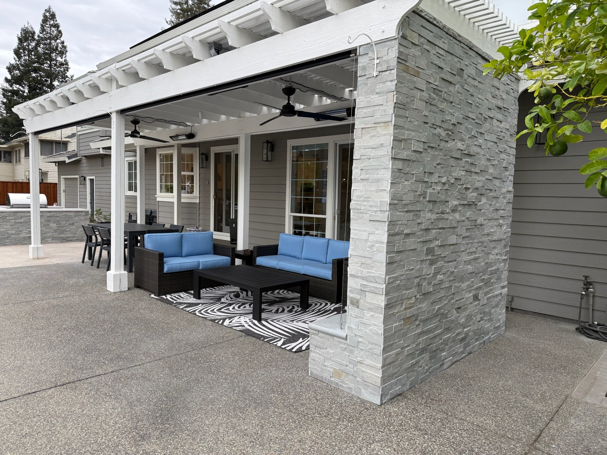 Backyard patio area with outdoor furniture, including a table and chairs, beneath a white pergola with ceiling fans, and a wall of gray stone and siding. There is a lemon tree with a lime or small green fruit on the right side of the image.