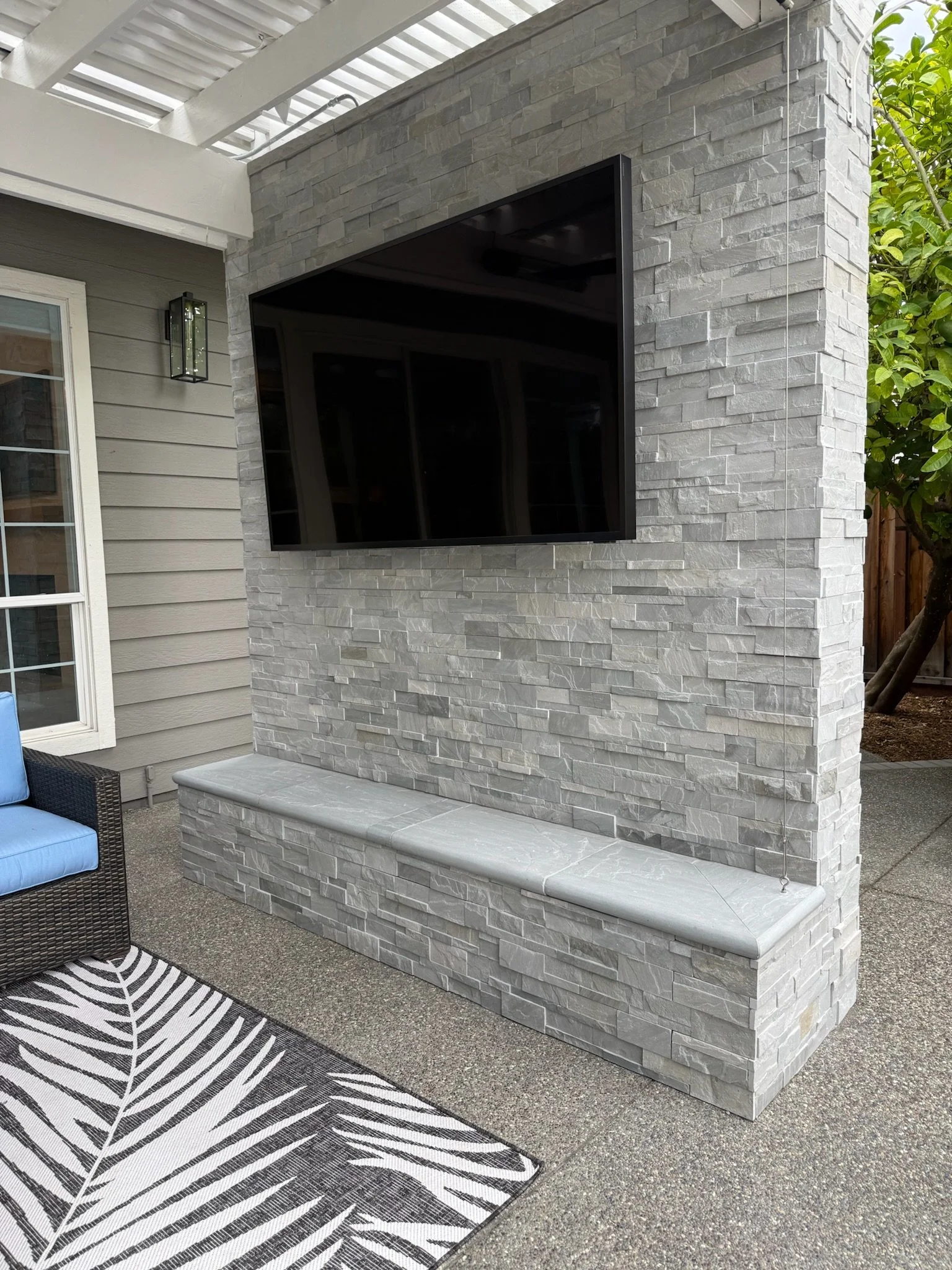 Exterior view of a house patio area featuring a stone wall with a mounted flat-screen TV, a gray and black patterned outdoor rug, and outdoor furniture including a wicker chair with a blue cushion.