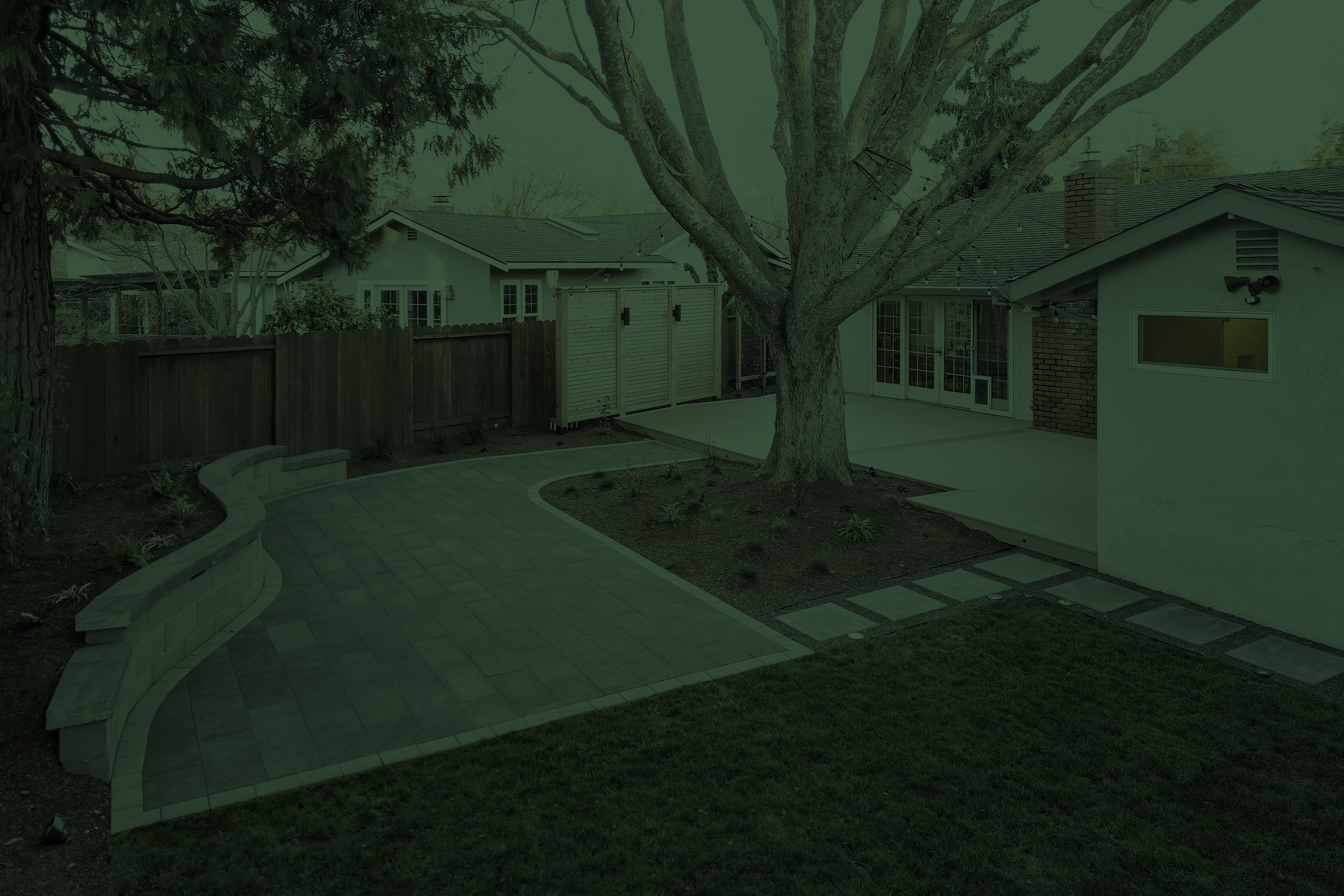 Backyard scene with large trees, a brick house with sliding glass doors, a wooden fence, a paved pathway, and outdoor lighting.