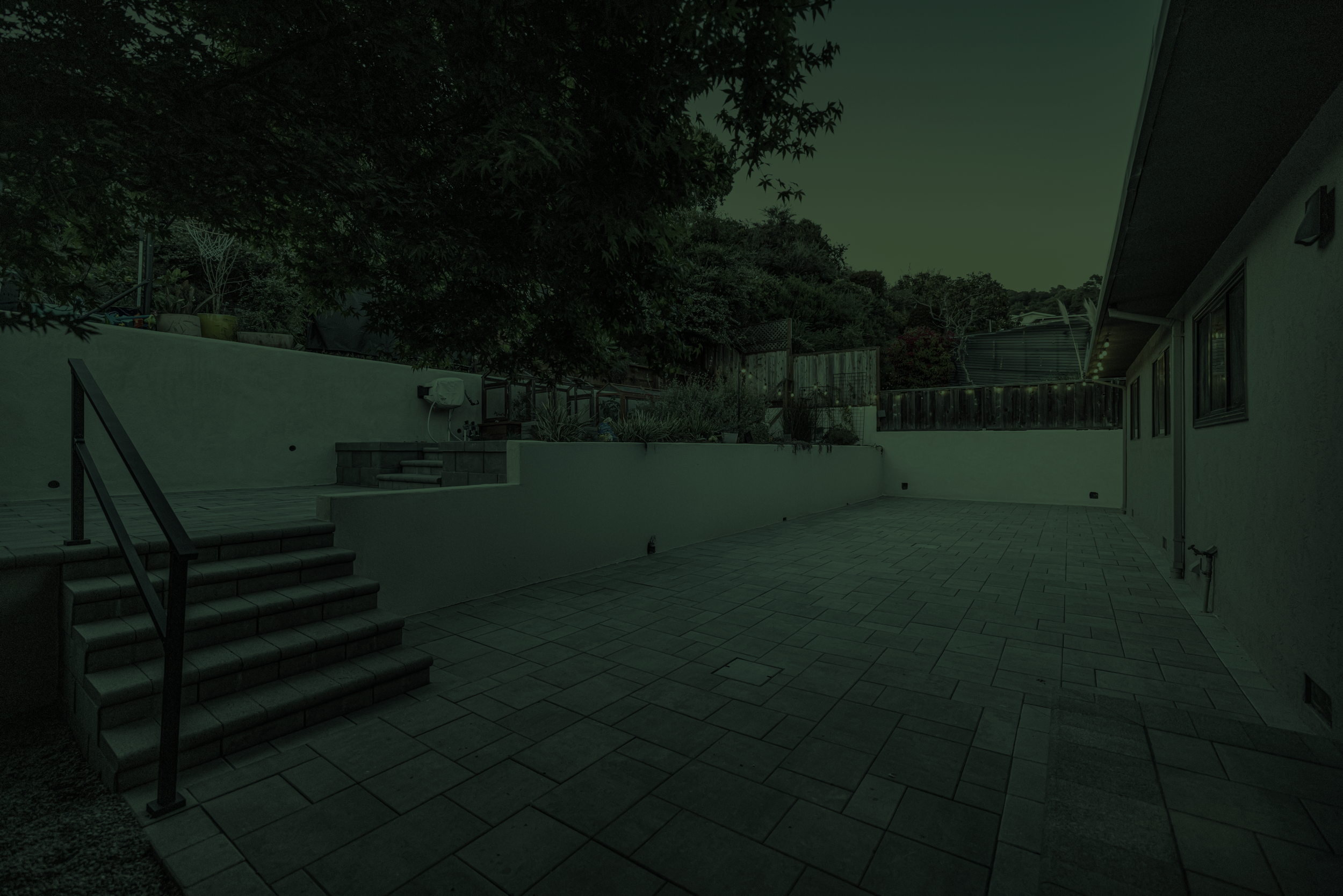 Backyard patio with stairs, fence, trees, and house, under a dark sky.