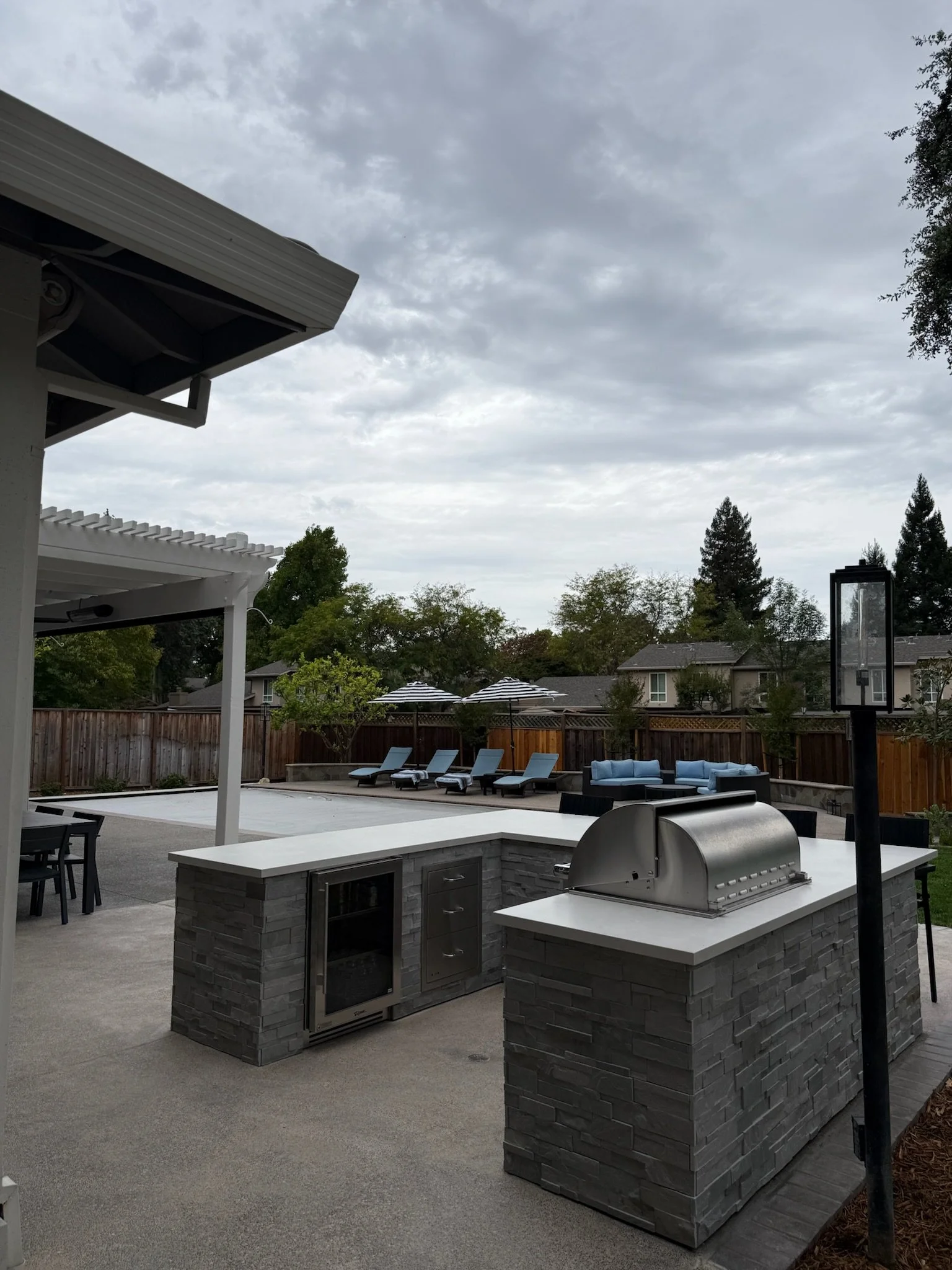 Backyard patio with built-in outdoor kitchen, grill, and seating area, surrounded by a wooden fence and trees under a cloudy sky.