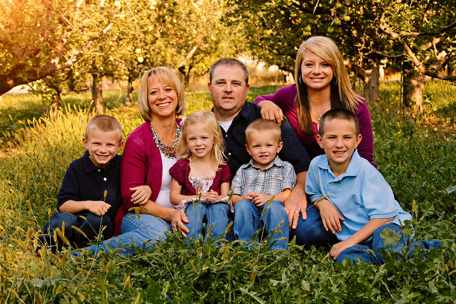 A family of seven sitting outdoors in a field of greenery and trees, smiling for the camera during sunset.