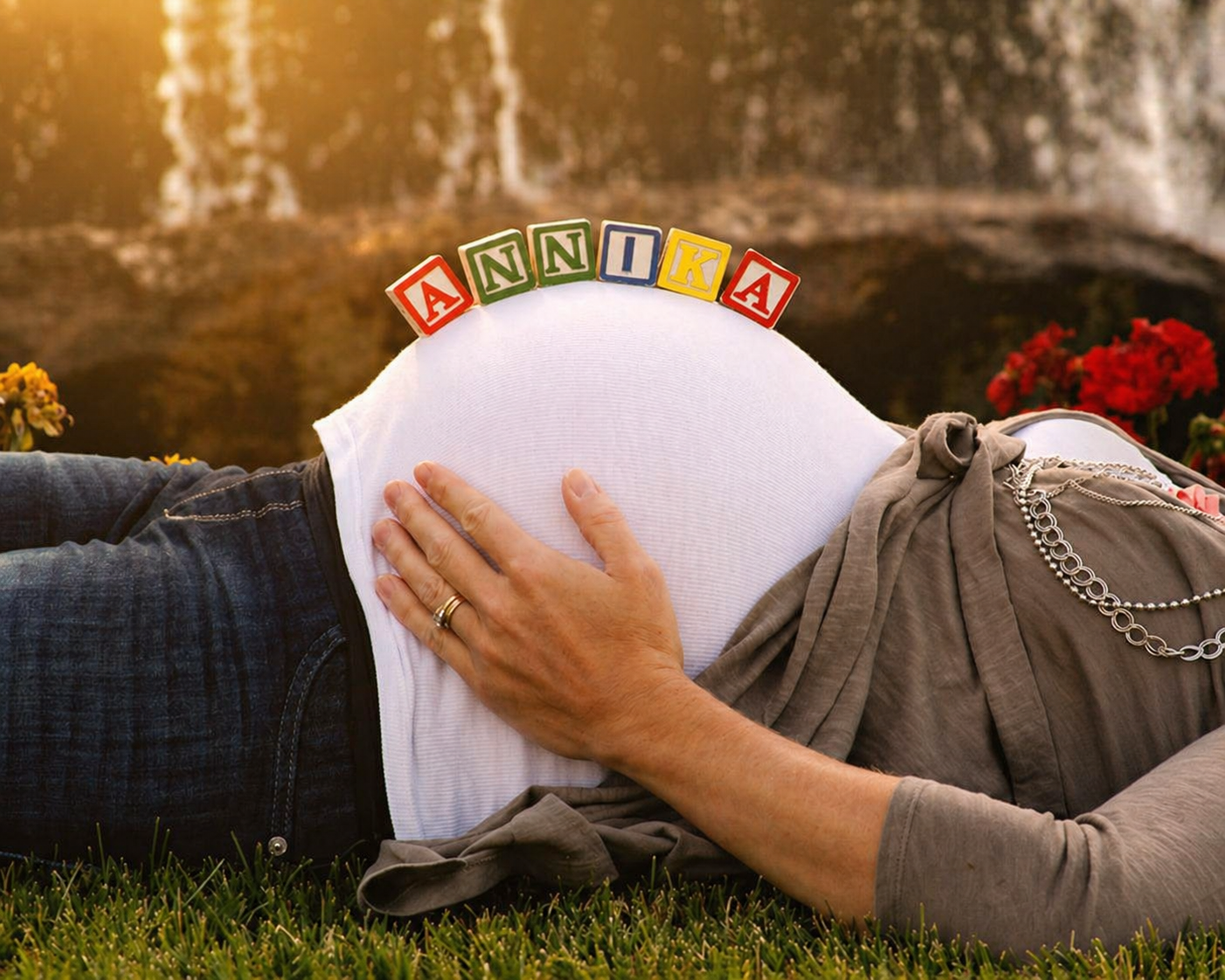 A pregnant woman lying on grass with her hand on her belly, decorated with colorful ABC blocks spelling 'ANNIKA' on her belly, outdoors during sunset with flowers in the background.