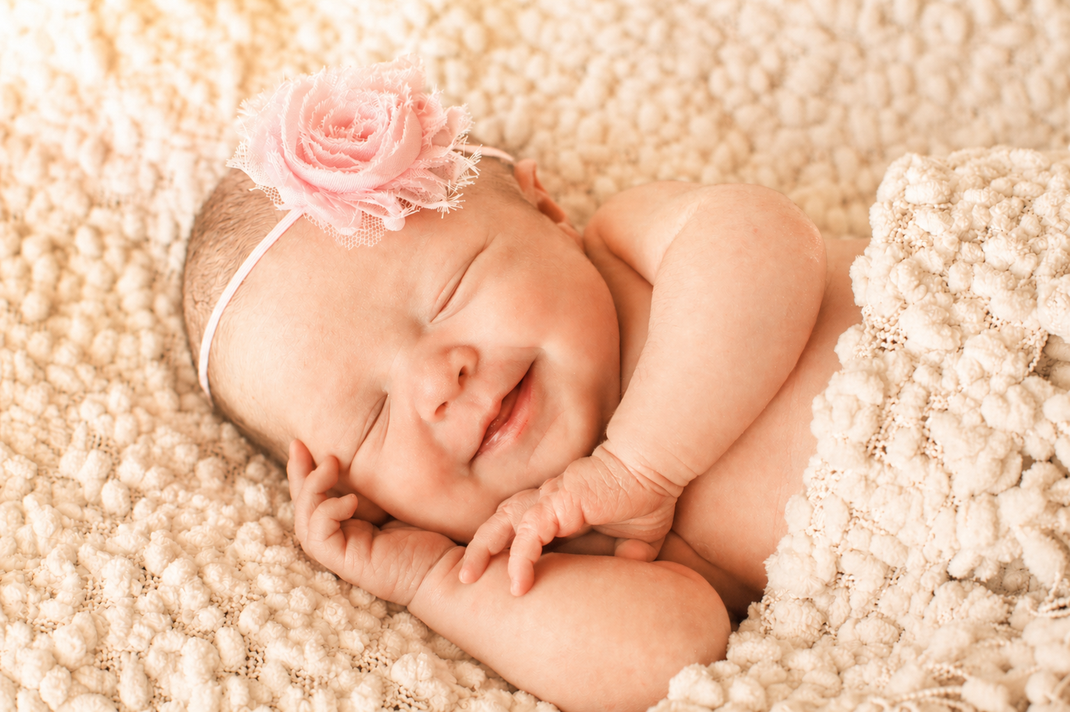 A smiling newborn baby girl, peacefully sleeping on a soft, textured cream-colored blanket, wearing a pink floral headband.