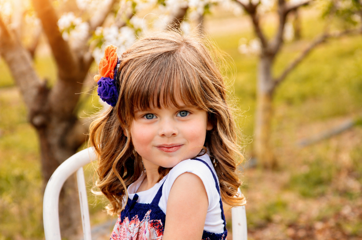 A young girl with wavy, light brown hair and blue eyes, wearing a floral dress with a white and navy top, sitting outdoors on a white metal chair in a garden or orchard, with trees and sunlight in the background.