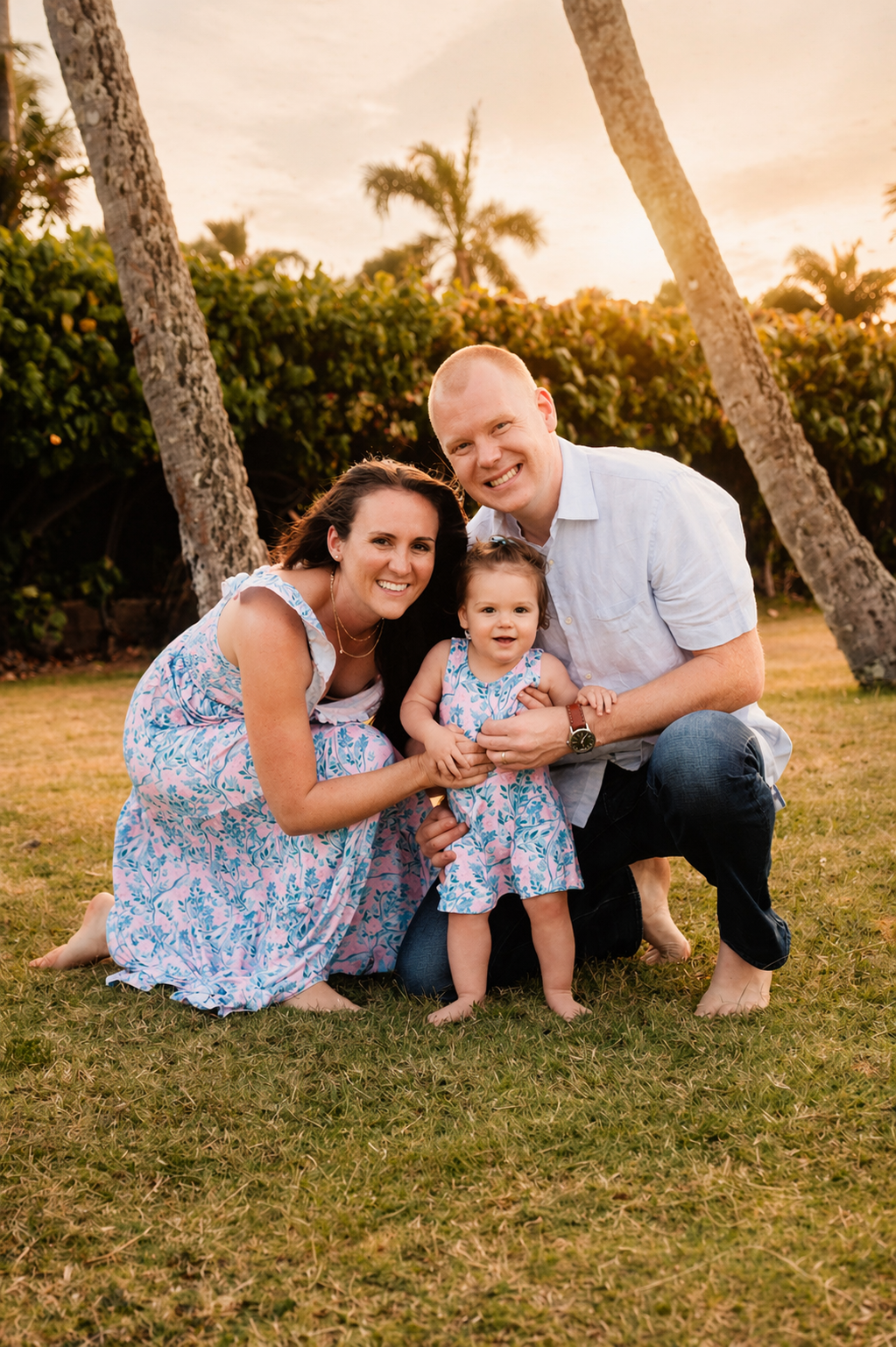 A happy family of three, a mother, father, and young girl, smiling and kneeling on a grassy area outdoors during sunset, with palm trees and lush greenery in the background.